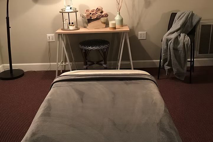 Massage table in a softly lit room with a side table, lamp, flowers, and a draped chair.