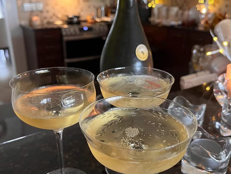 Three champagne coupes with pale sparkling drink on a table, with a bottle and blurred lights in the background