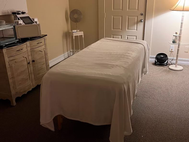 Massage table in a softly lit treatment room with a lamp, fan, and cabinet.