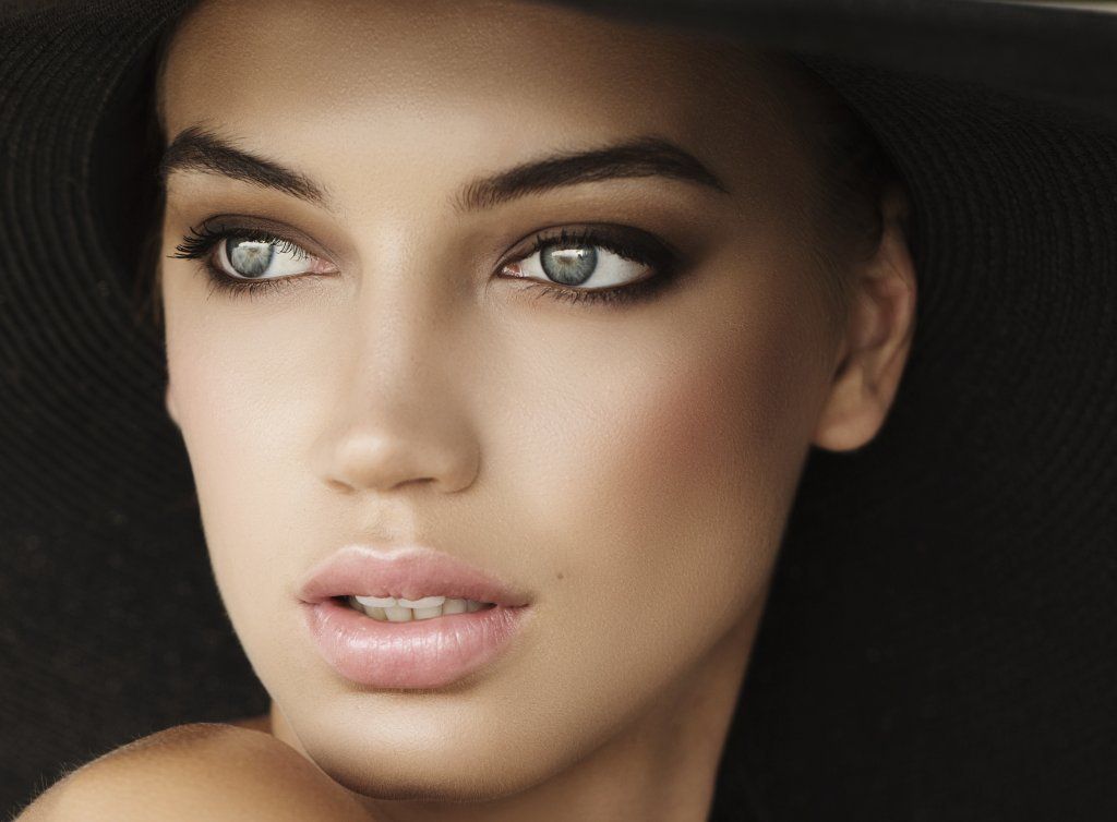 Woman with smoky eye makeup, wearing a black hat, looking left, soft lighting.