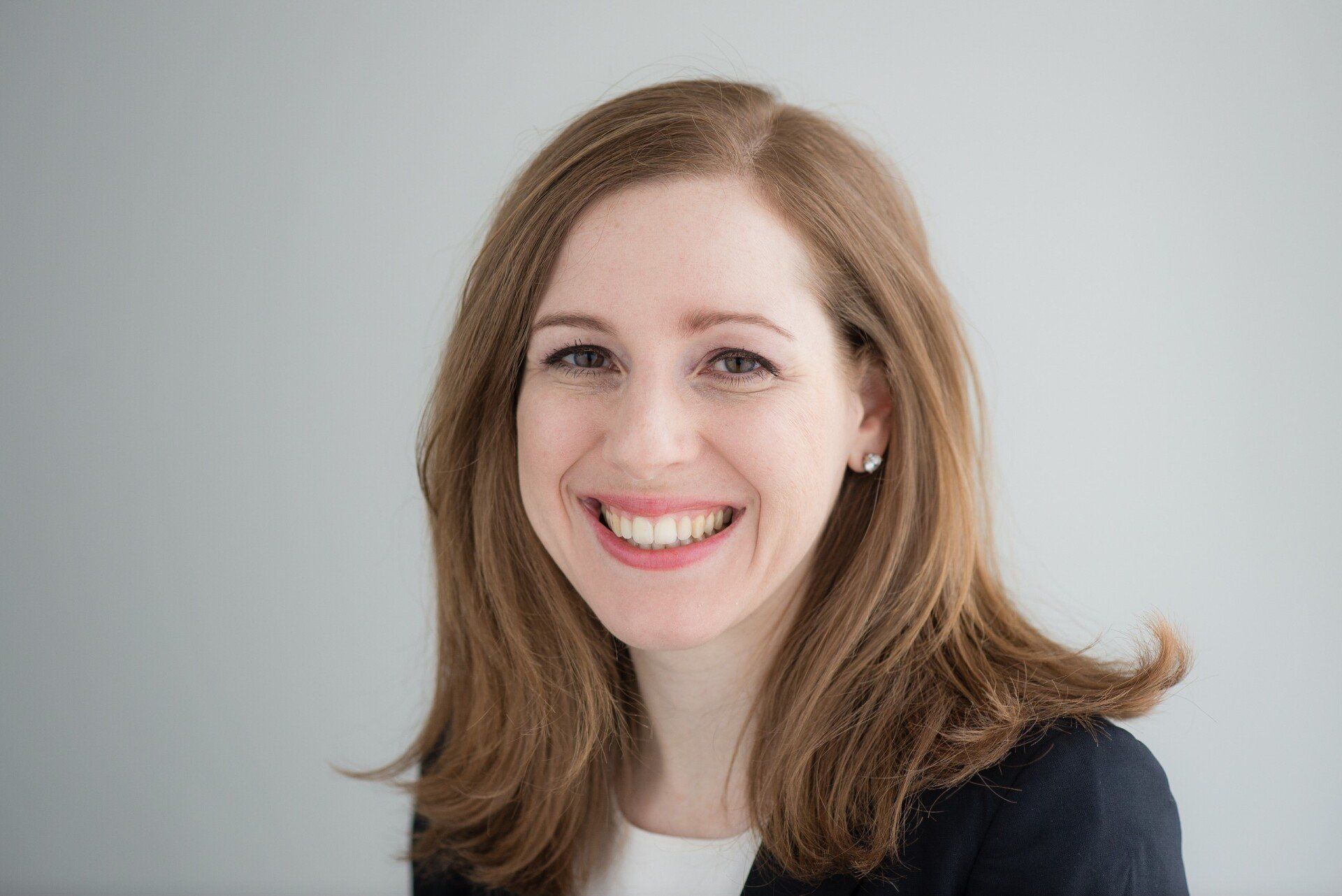 Woman with long brown hair smiles at the camera, wearing a navy blazer and white top, against a grey background.