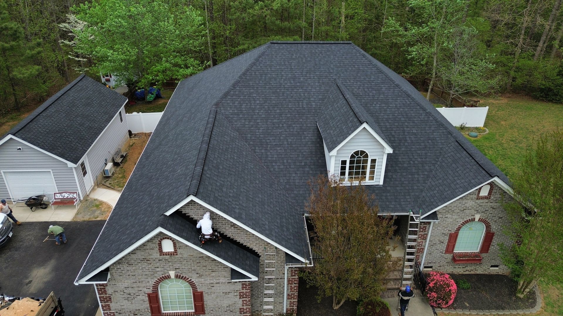 Aerial view of a large gray-roofed house with dormer windows, front yard, and detached garage.