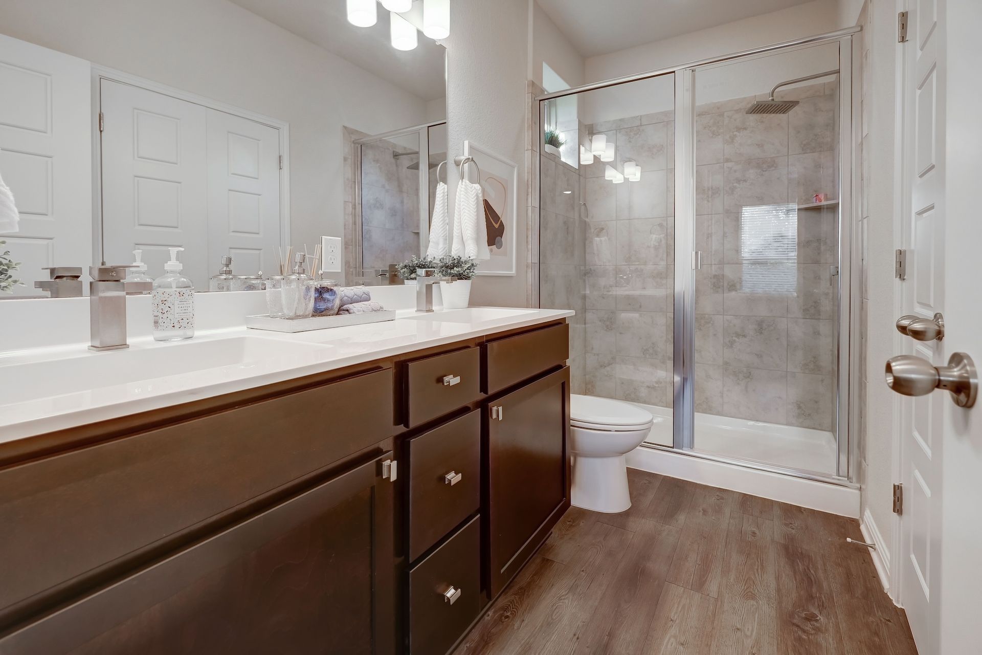 Bathroom with dark brown vanity, double sinks, glass shower, and wood-look flooring.
