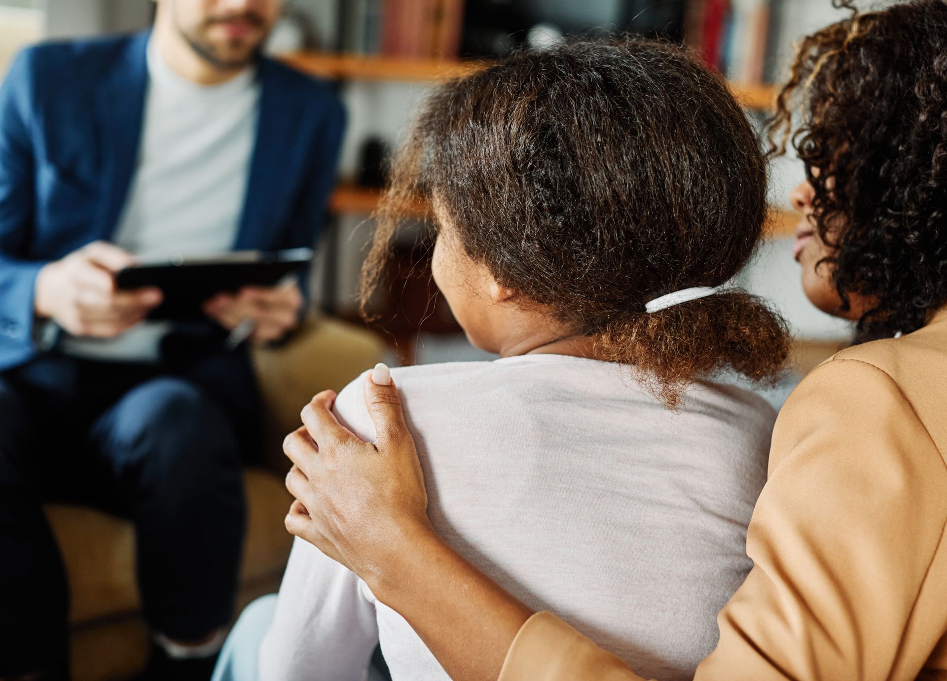 A woman is hugging a little girl while a man holds a tablet.