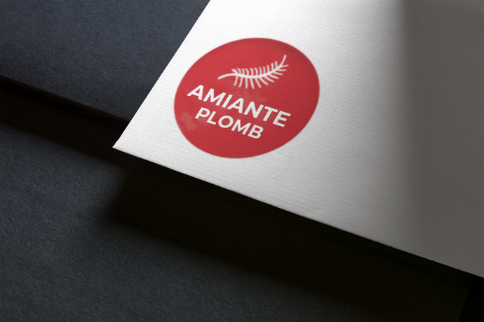 Cercle rouge avec le texte « AMIANTE PLOMB » et un motif de feuille sur papier blanc, appuyé sur une surface sombre.