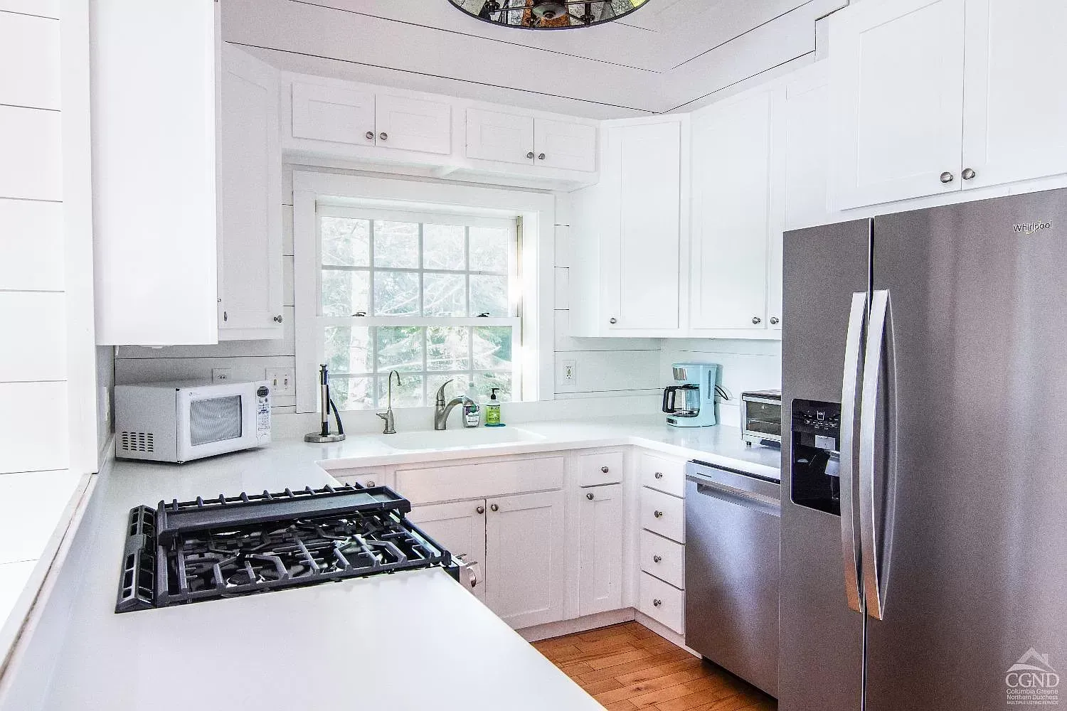 A kitchen with white cabinets , a stove , a refrigerator , a microwave , and a window.
