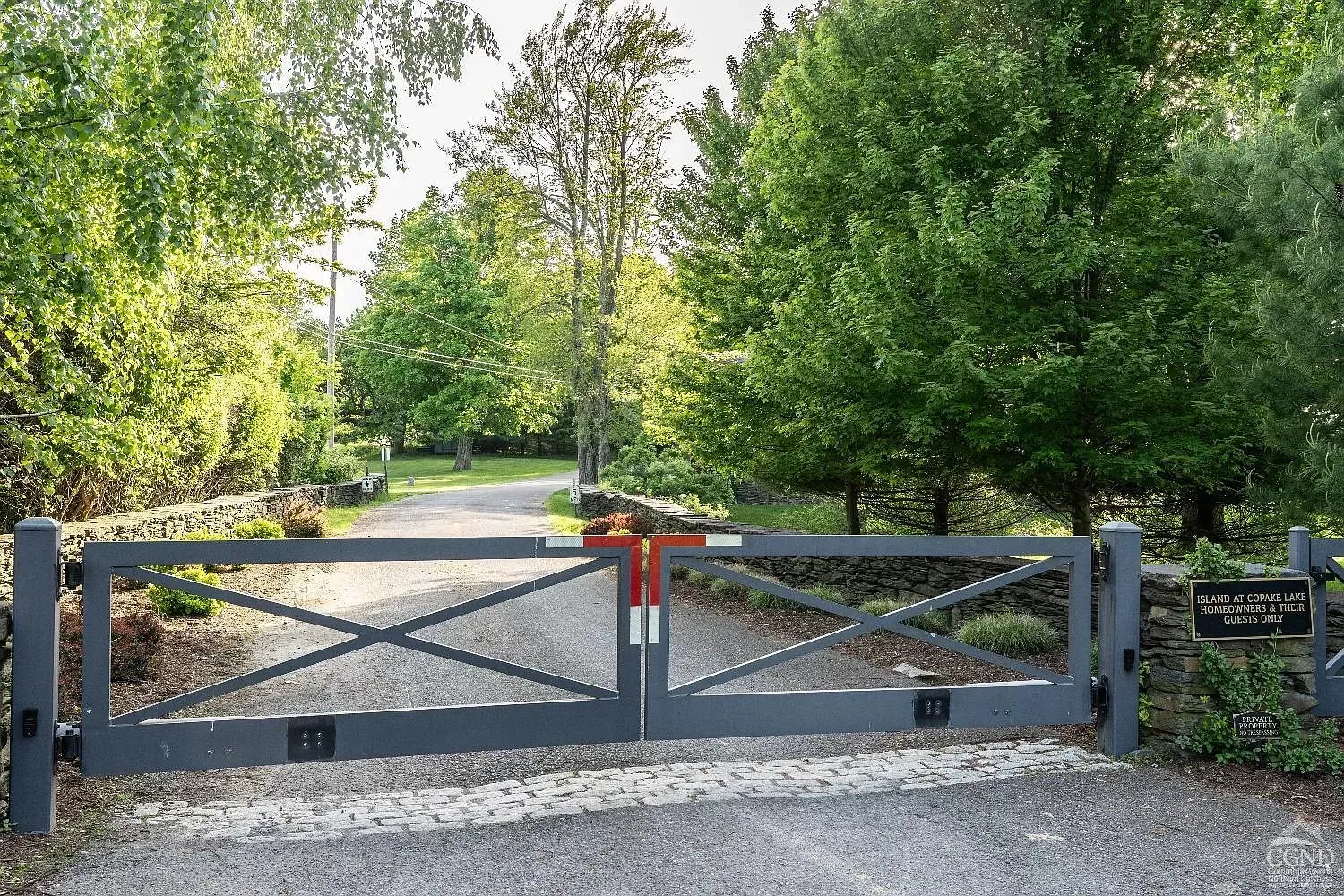 A large metal gate is closed on a gravel road surrounded by trees.