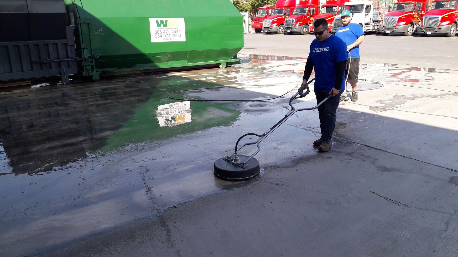 Man pressure washing a concrete surface outdoors near a green dumpster and trucks.