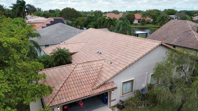 Aerial view of a house with orange tile roof, stucco walls, surrounded by green trees and other houses.