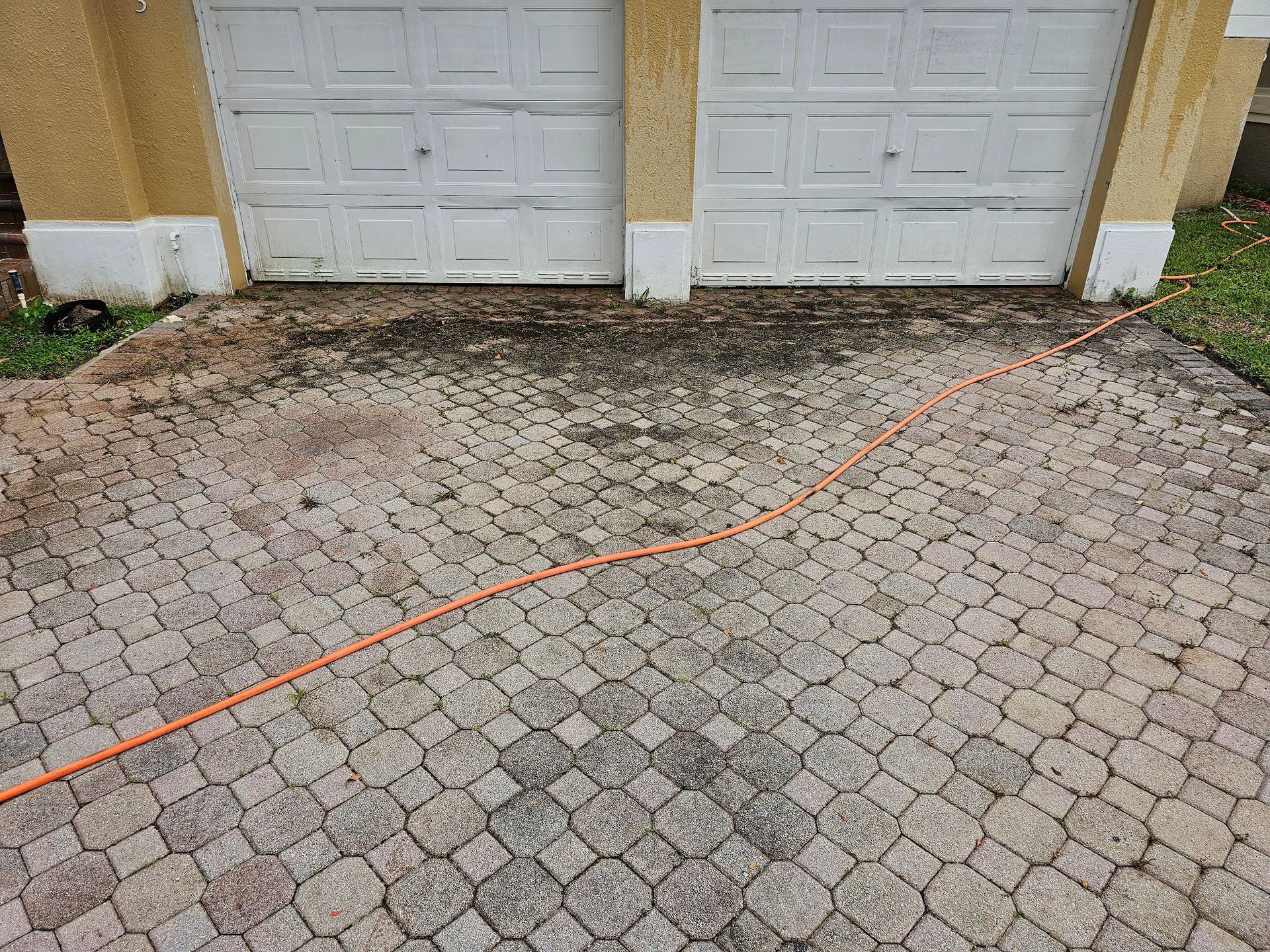 Driveway with orange hose, leading towards two garage doors. The driveway has patterned, dark patches.