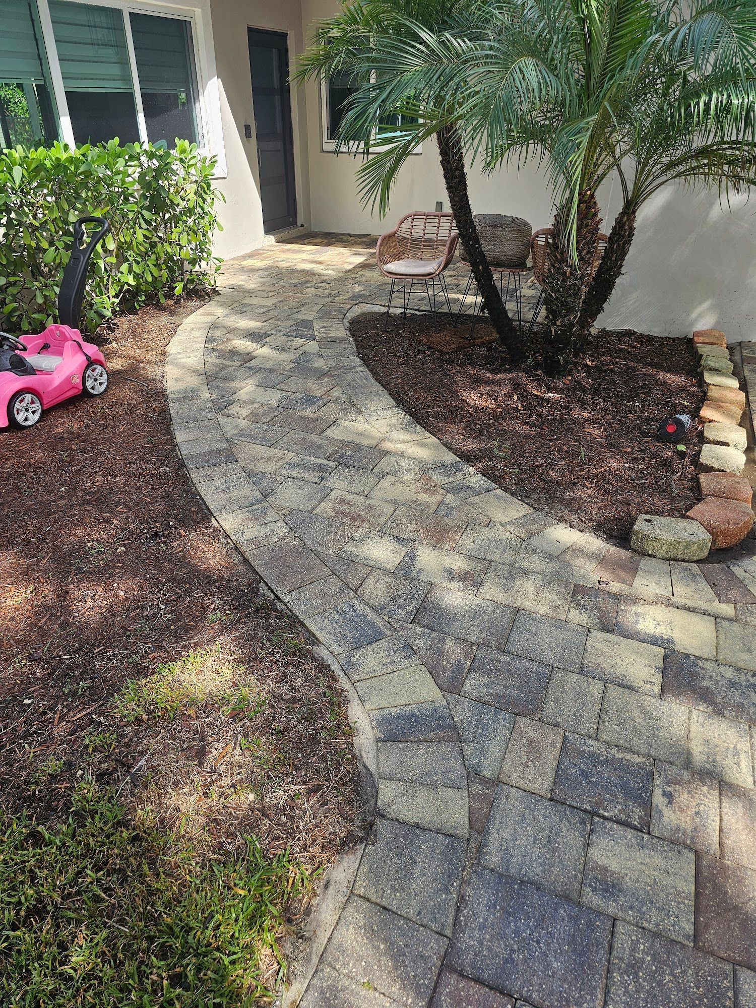 Brick pathway curves towards a front door, next to a manicured bed and palm tree.