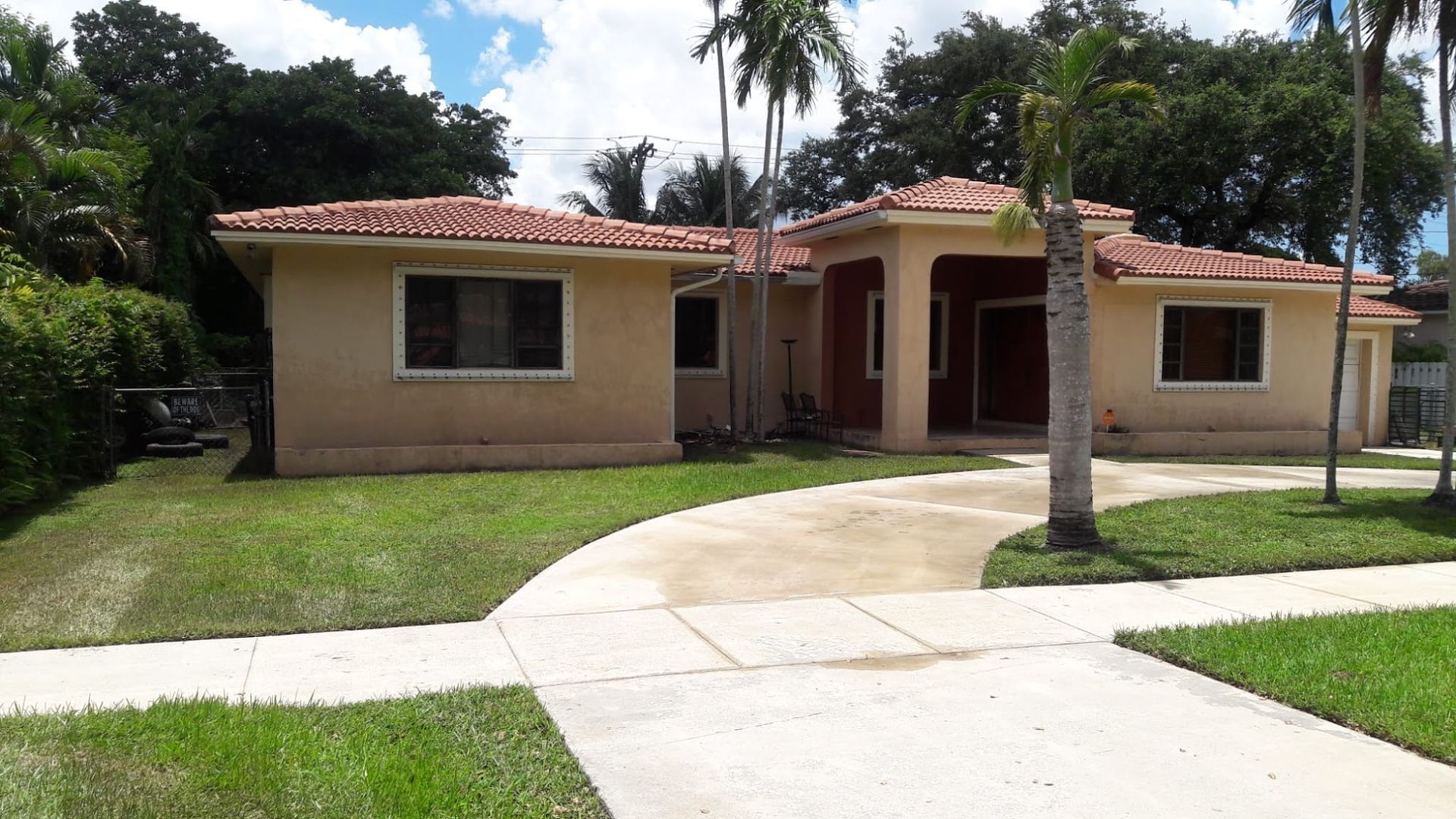 Tan stucco house with orange tile roof and curved driveway, front yard with green grass, blue sky.