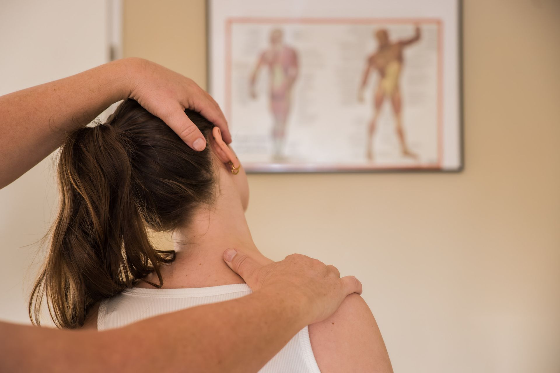 Handen onderzoeken de nek en schouder van een persoon. Op de achtergrond is een poster van de menselijke anatomie te zien.