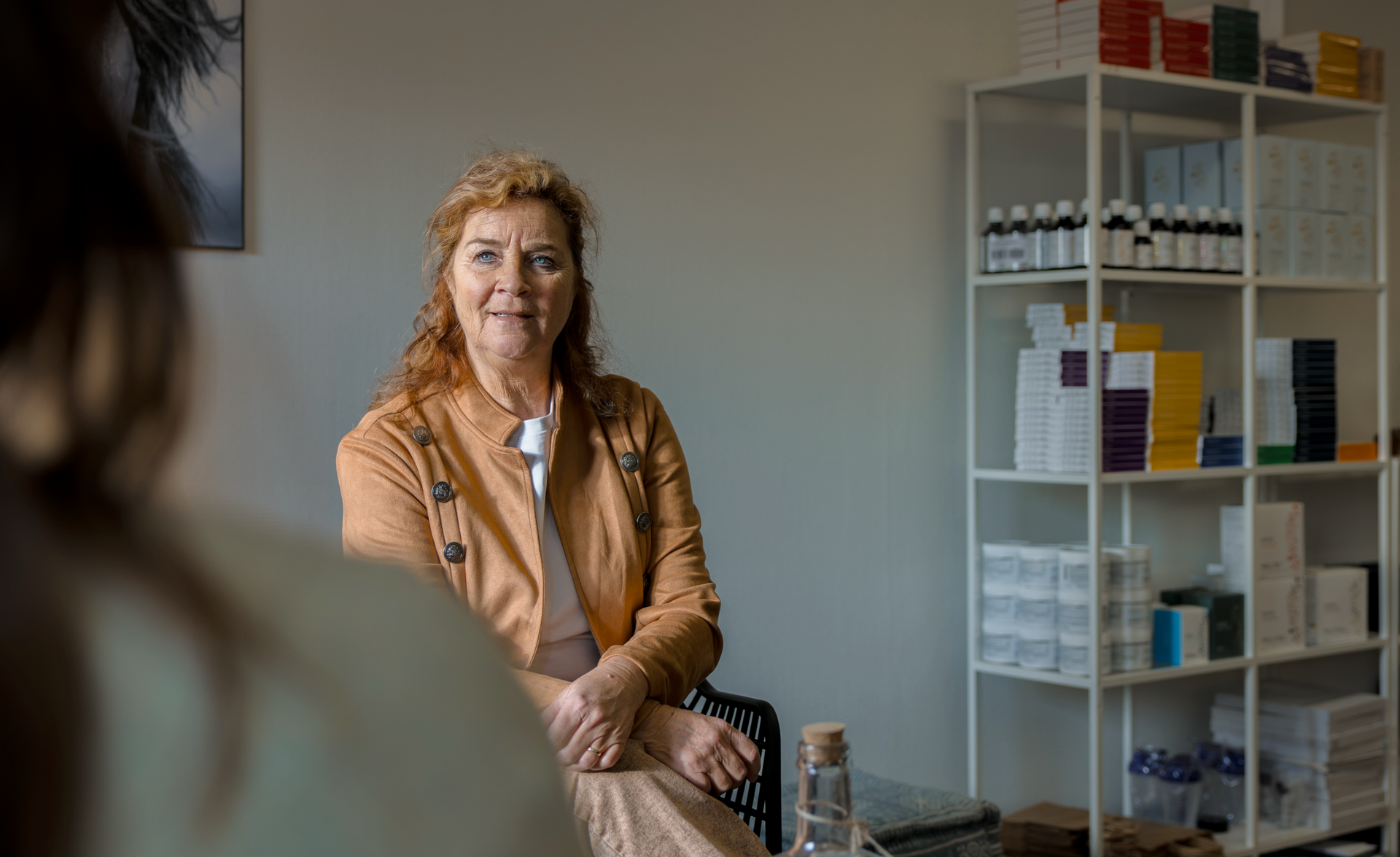 Een vrouw in een oranje vest zit tegenover een persoon en kijkt aandachtig. Achter haar staan witte schappen met producten.