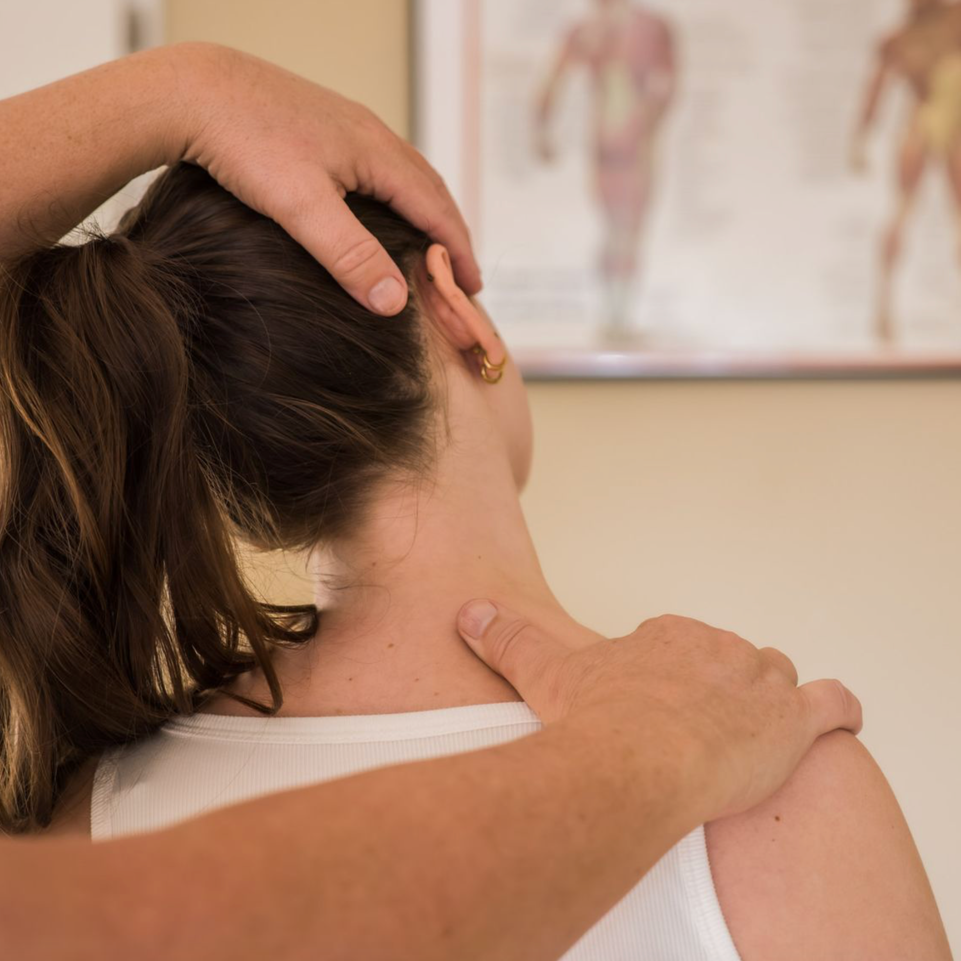 Handen die de nek en schouder van een persoon manipuleren. Een zorgomgeving met een anatomiekaart op de achtergrond.
