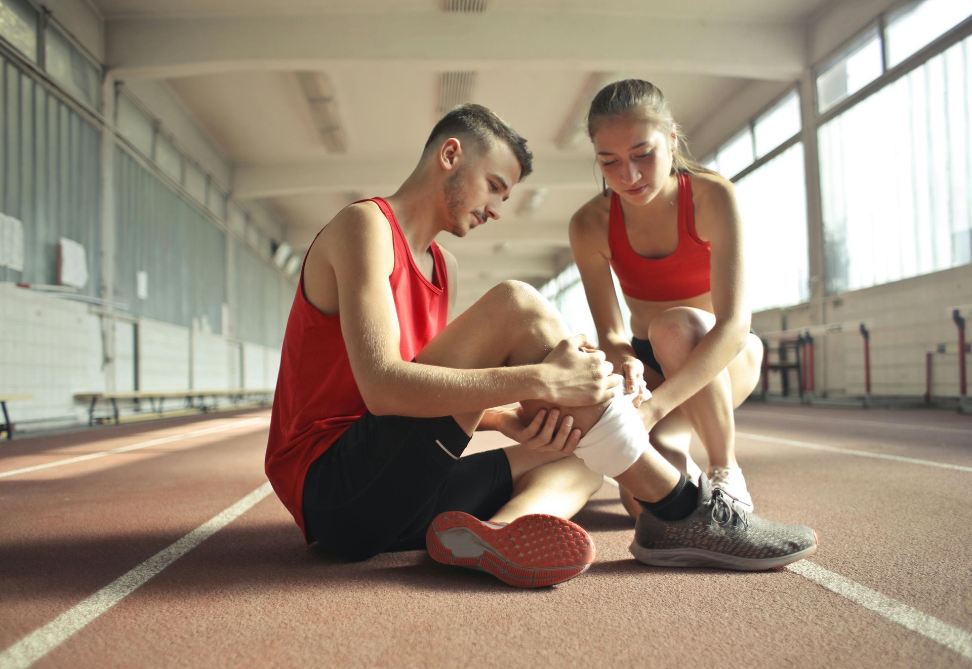 Een atleet met een verbonden been wordt op de atletiekbaan geholpen door een andere atleet. Hij draagt rode kleding.