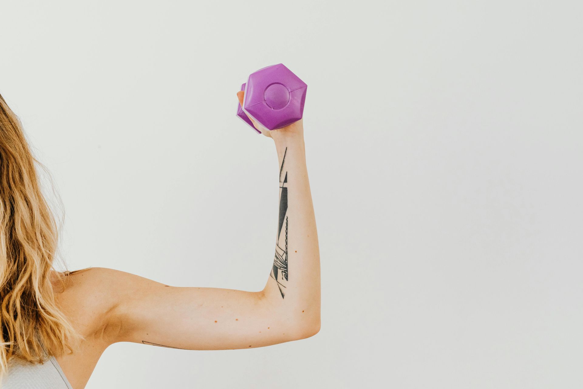 Vrouw tilt een paarse halter op, met gestrekte arm, waarbij een tatoeage zichtbaar is. Witte achtergrond.