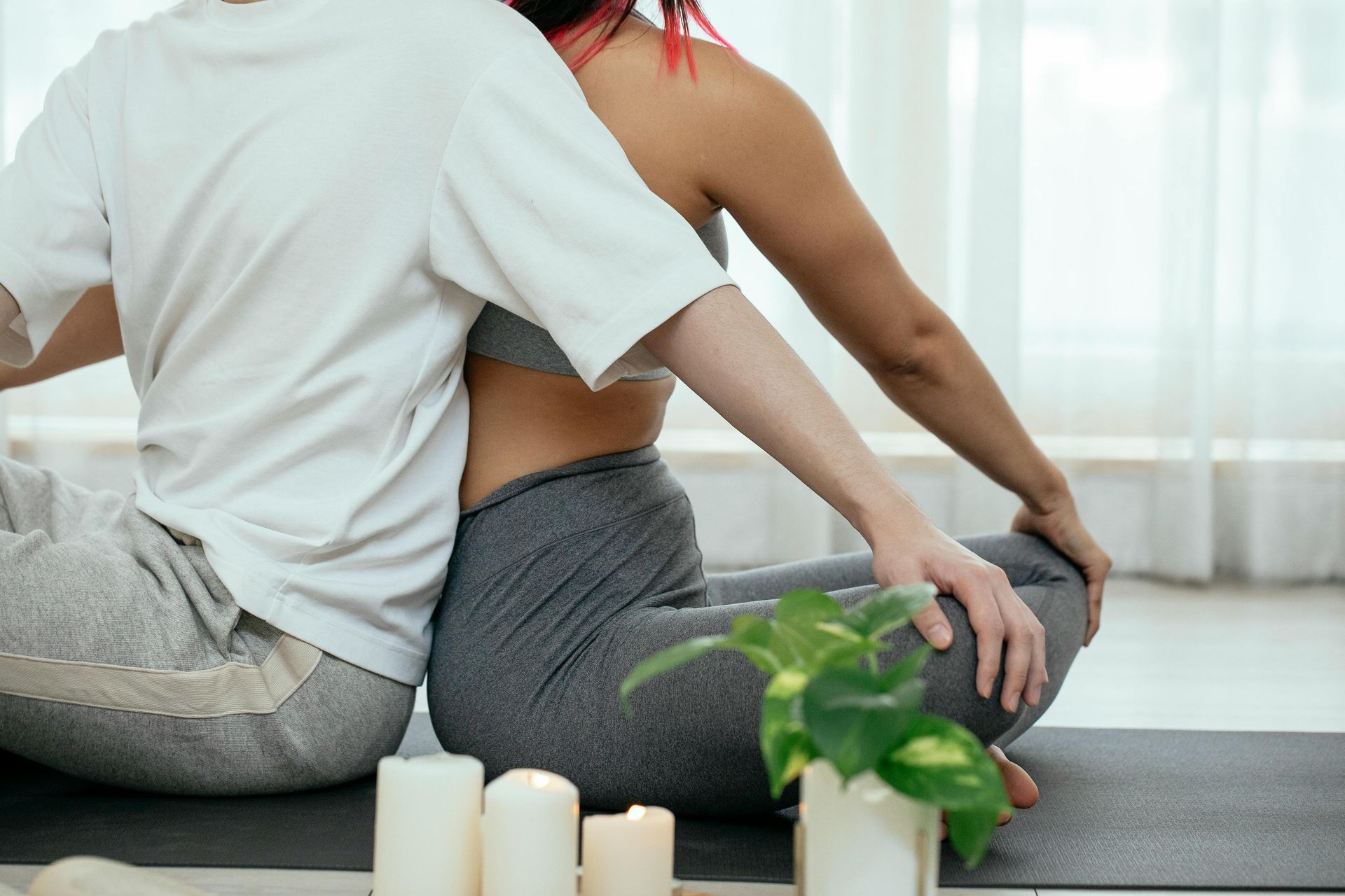 Two people seated on a mat, one in a white shirt, the other in a gray sports bra, touching hands, with candles.