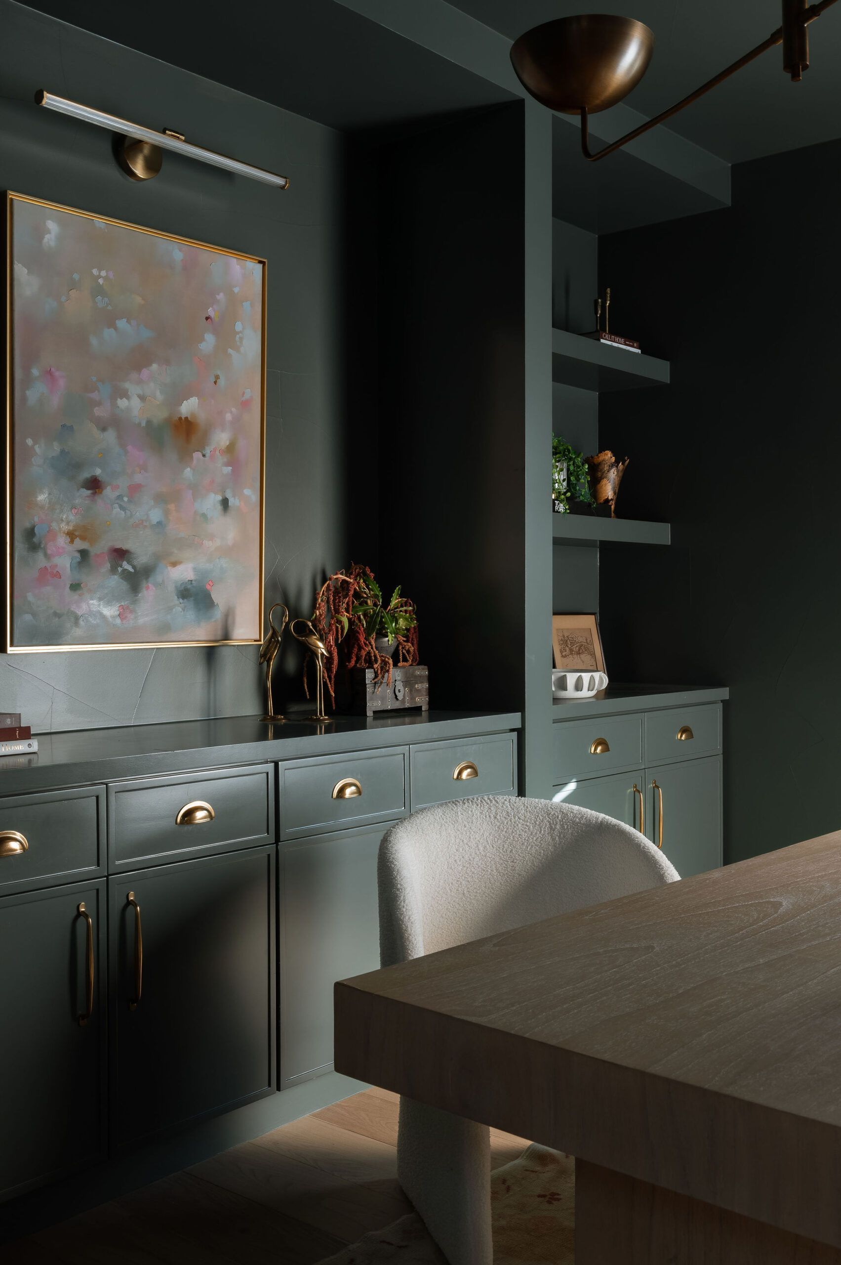 A dark, moody home office with olive green cabinetry, gold hardware, open shelving, abstract art, and a plush white chair.