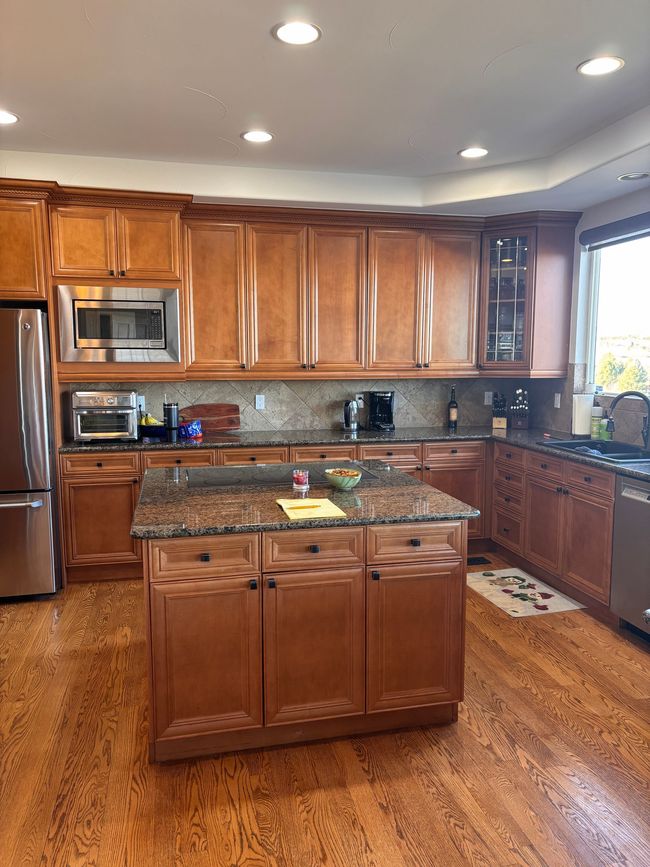 A wooden-cabinet kitchen featuring a granite island, stainless steel appliances, and hardwood floors.