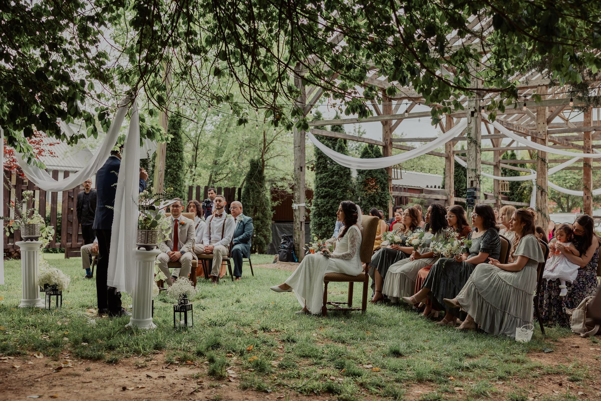 Wide view of an outdoor ceremony with guests and wedding party seated under the pergola