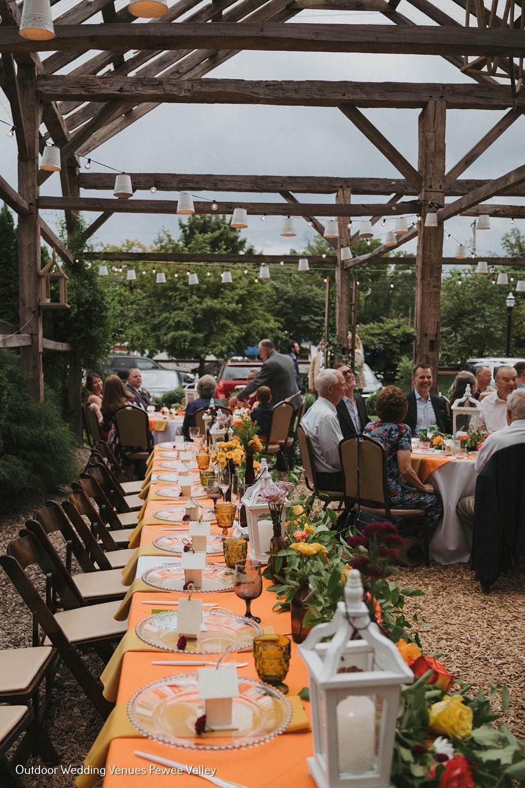Outdoor wedding reception with long flower-lined table under wooden pergola, guests seated and dining
