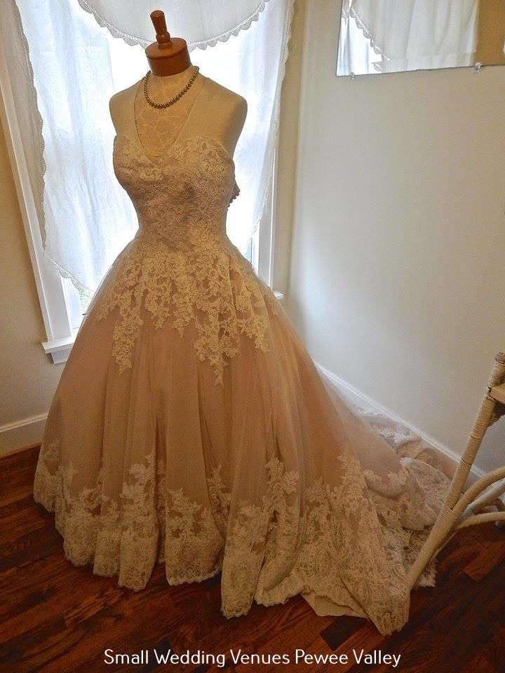 Beige lace ball gown displayed on a mannequin in a softly lit room with white curtains.