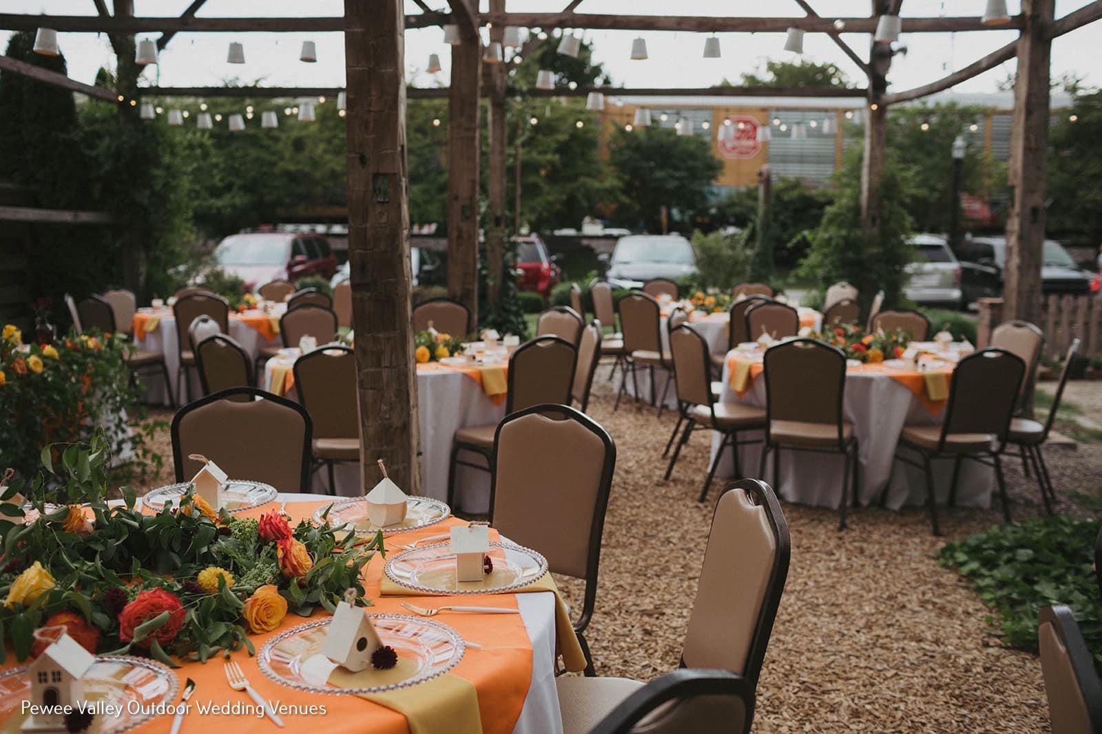 Outdoor wedding reception with round tables, orange tablecloths, and floral centerpieces under a wooden pergola.