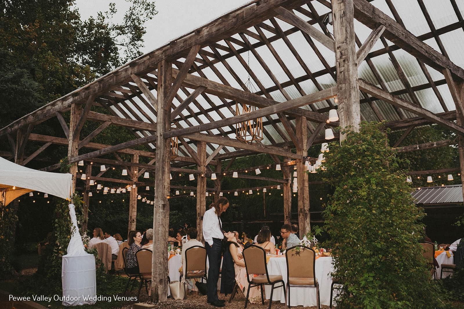 Outdoor wedding reception under a wooden pavilion with string lights and seated guests at round tables