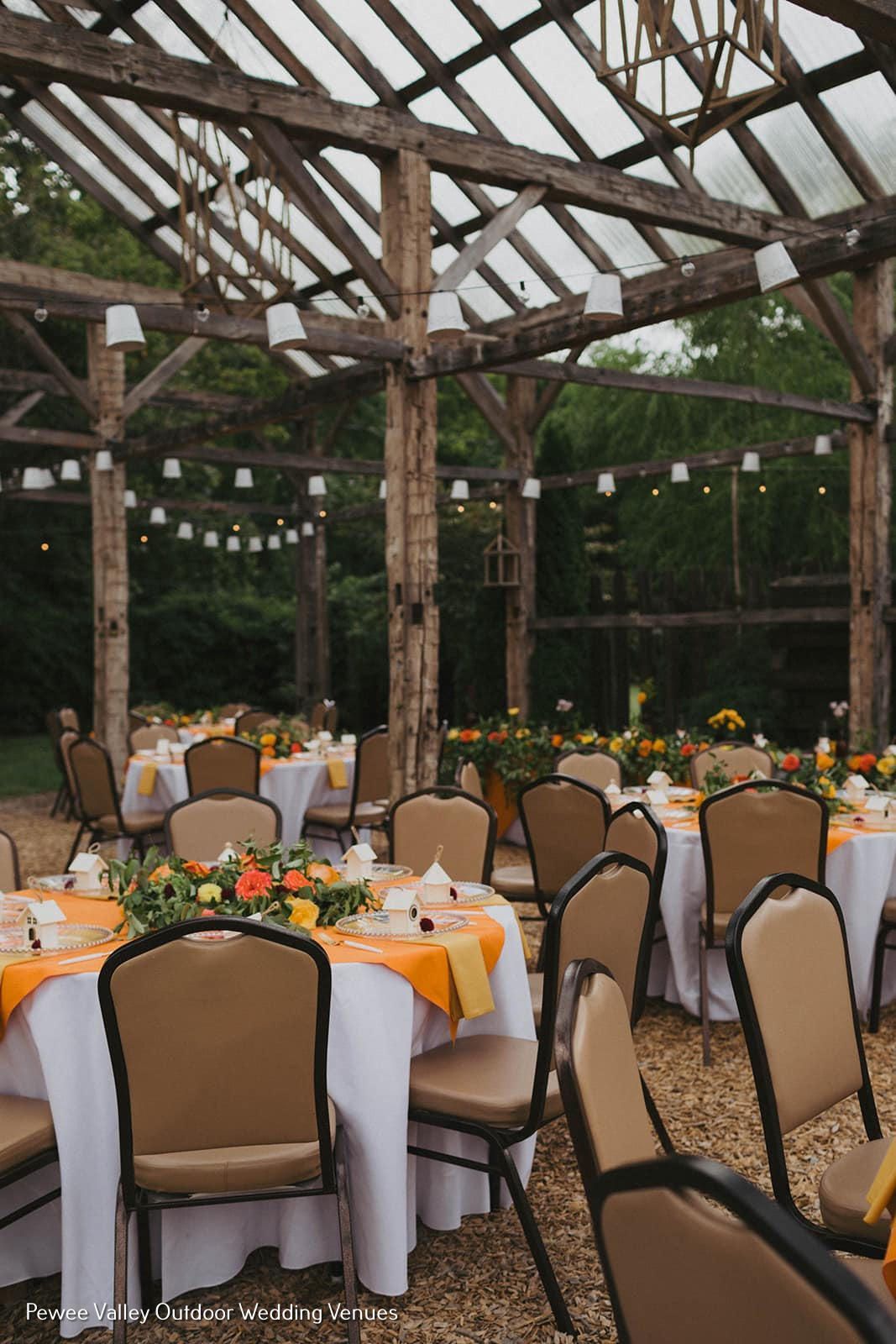 Outdoor banquet under a wooden pavilion with round tables, white linens, orange napkins, and string lights