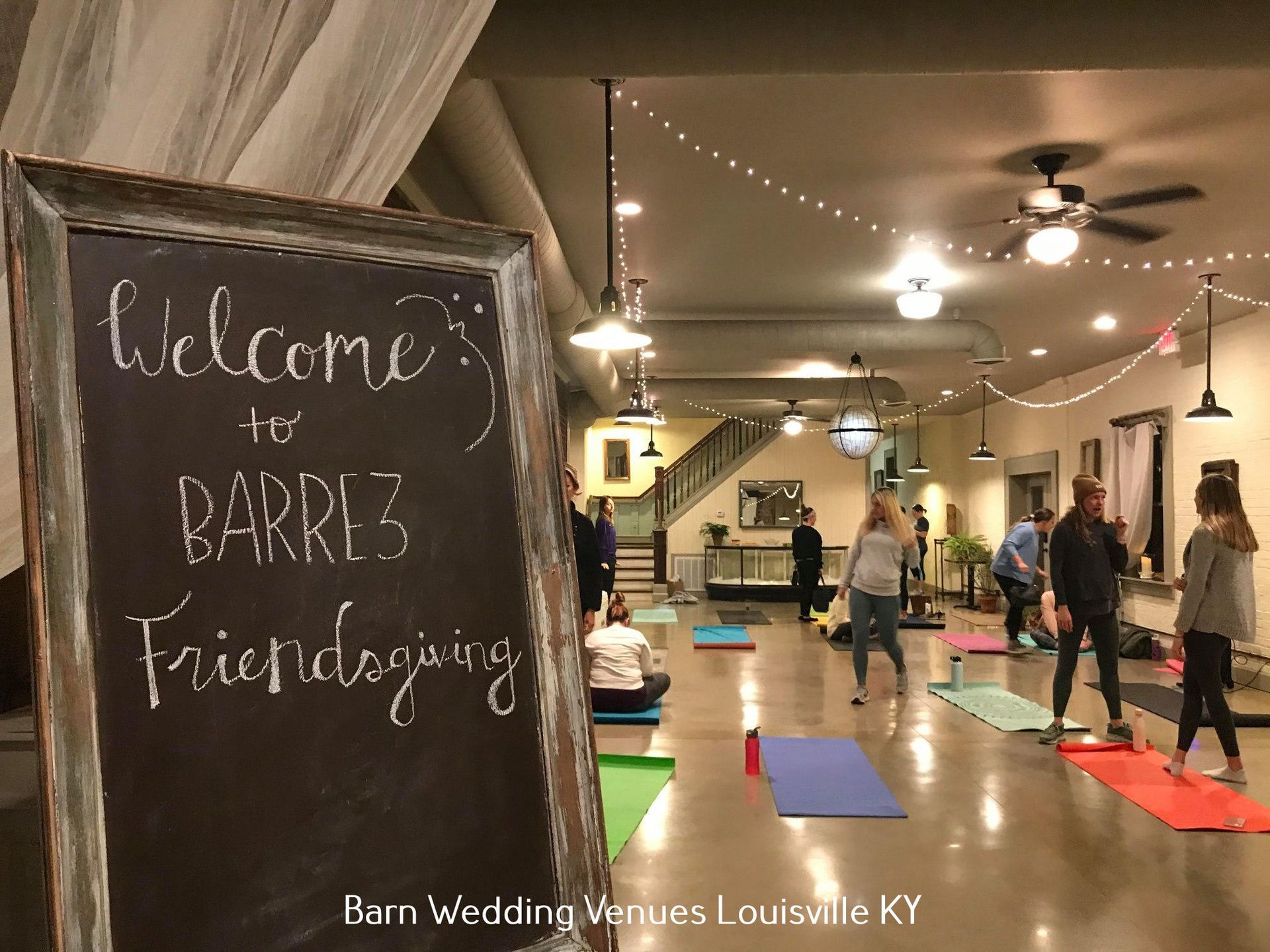 Chalkboard “Welcome to” sign at a wedding venue, with guests in a warmly lit rustic hall.