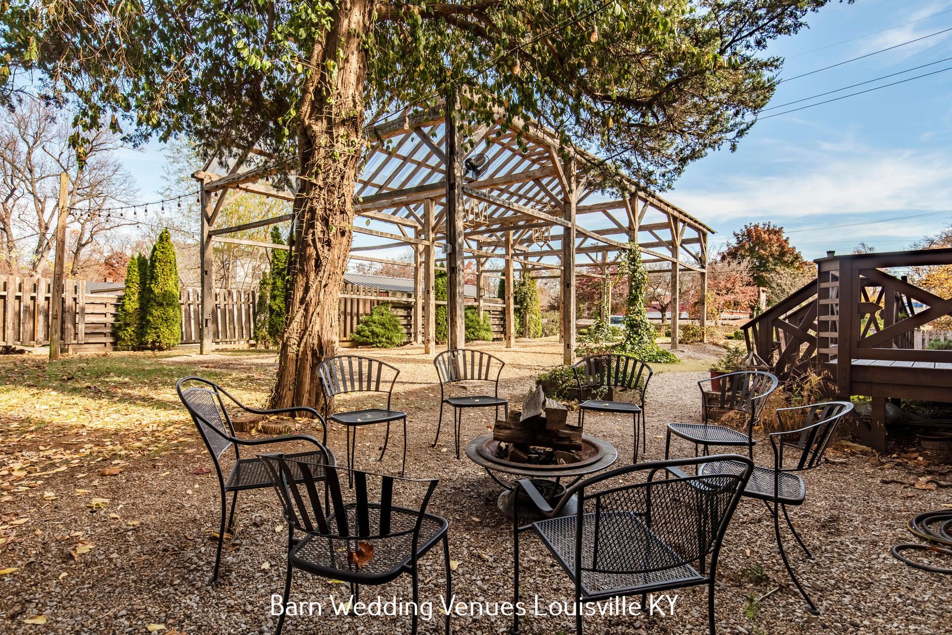 Outdoor patio with metal chairs and tables under trees beside a wooden pavilion on a sunny day