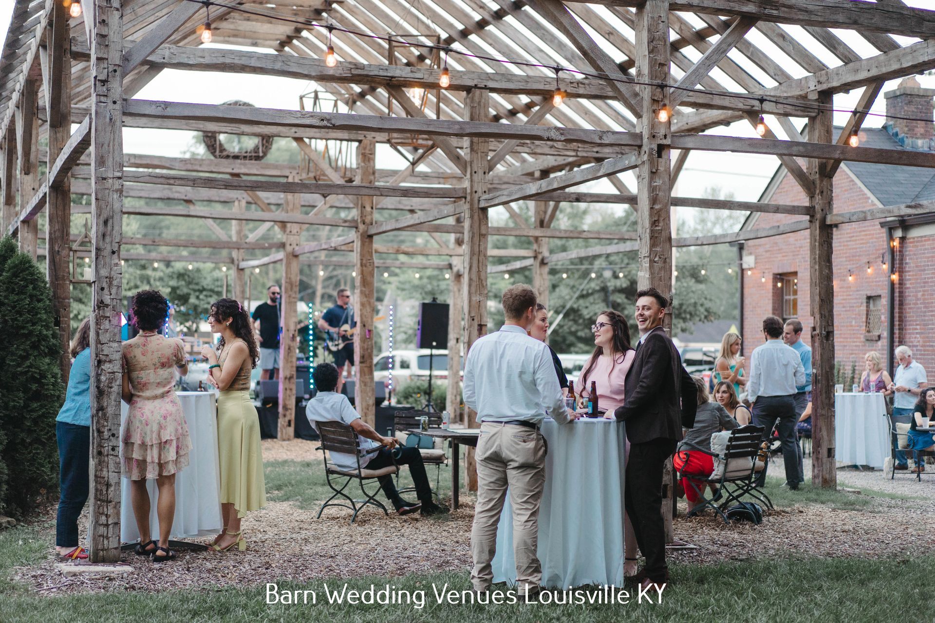 Guests mingling under a wooden pavilion at an outdoor wedding venue, with tables and chairs nearby.