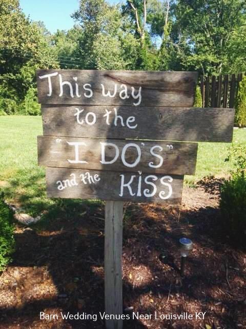 Wooden wedding sign reads “This way to the I dos and the kiss” in a grassy outdoor setting.