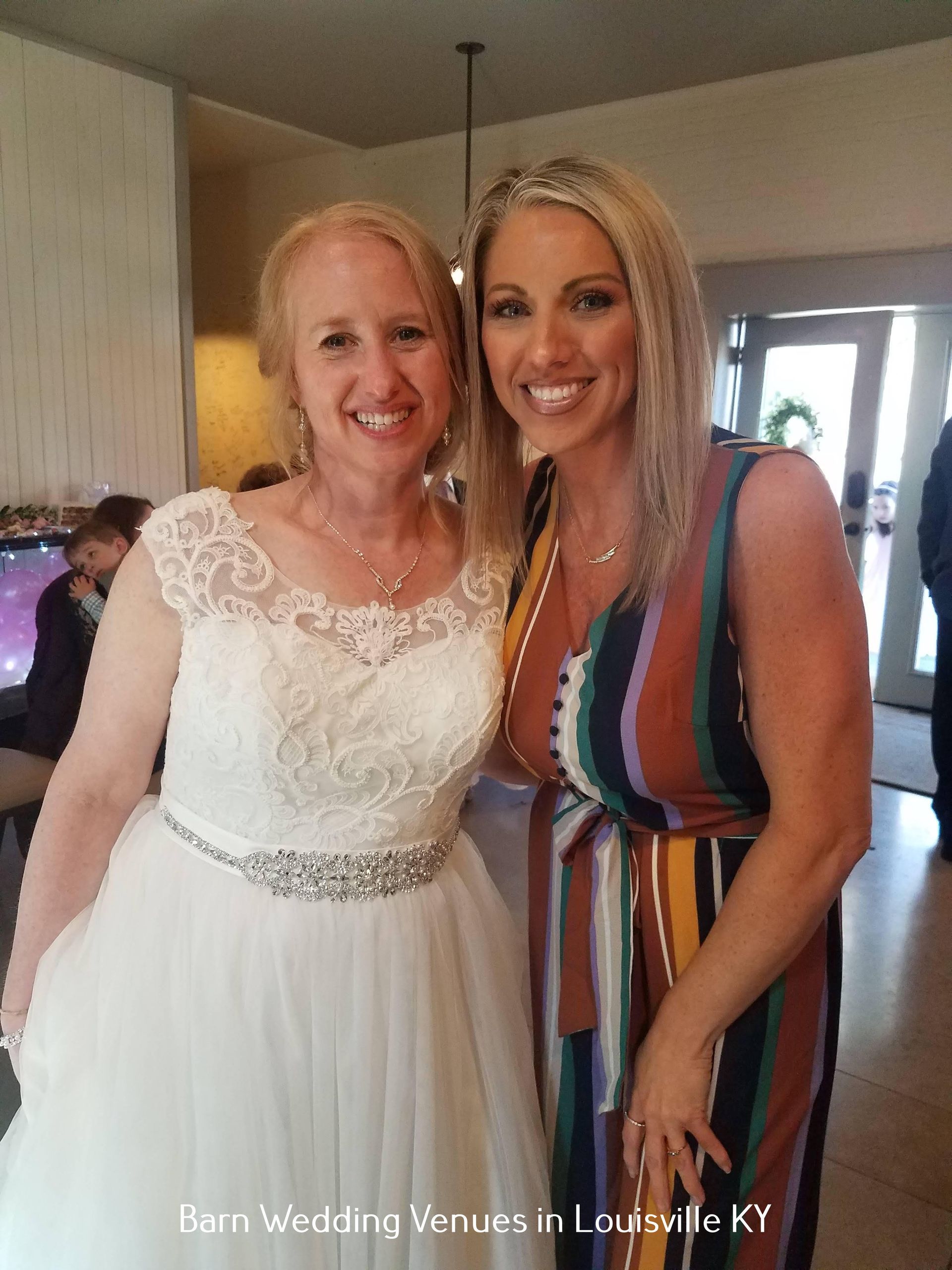 Two women posing indoors at a wedding venue, one in a white gown and one in a striped dress.