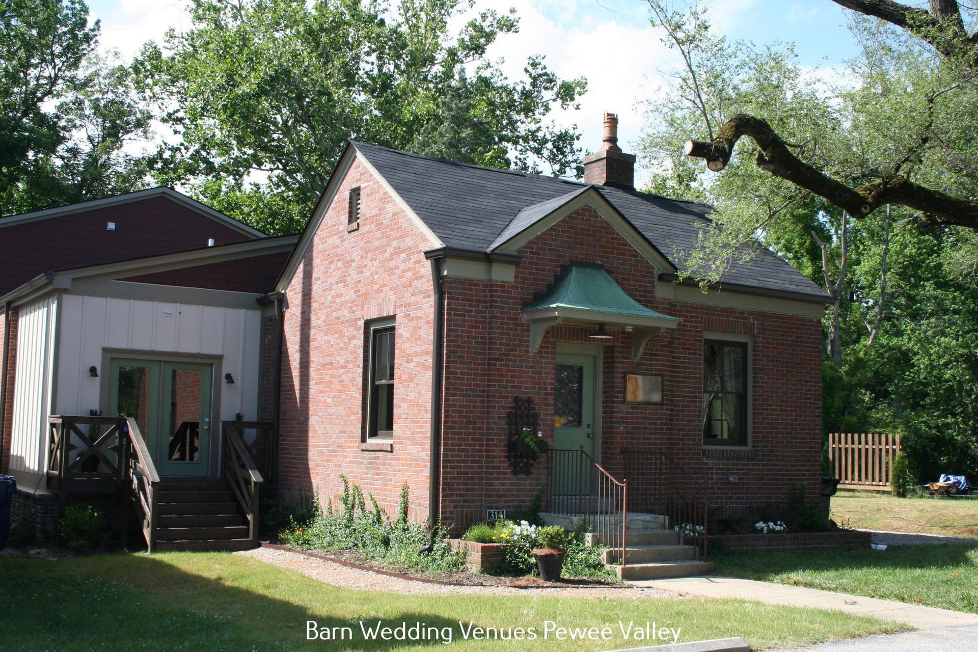 Brick house with green awning and porch, beside a larger dark building on a sunny tree-lined street