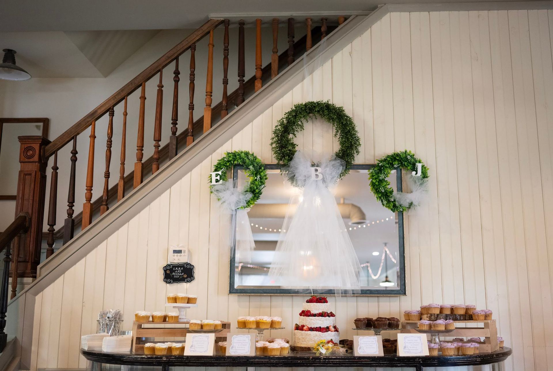 Decorated dessert table beneath a staircase with greenery, white drapery, and a cake centerpiece