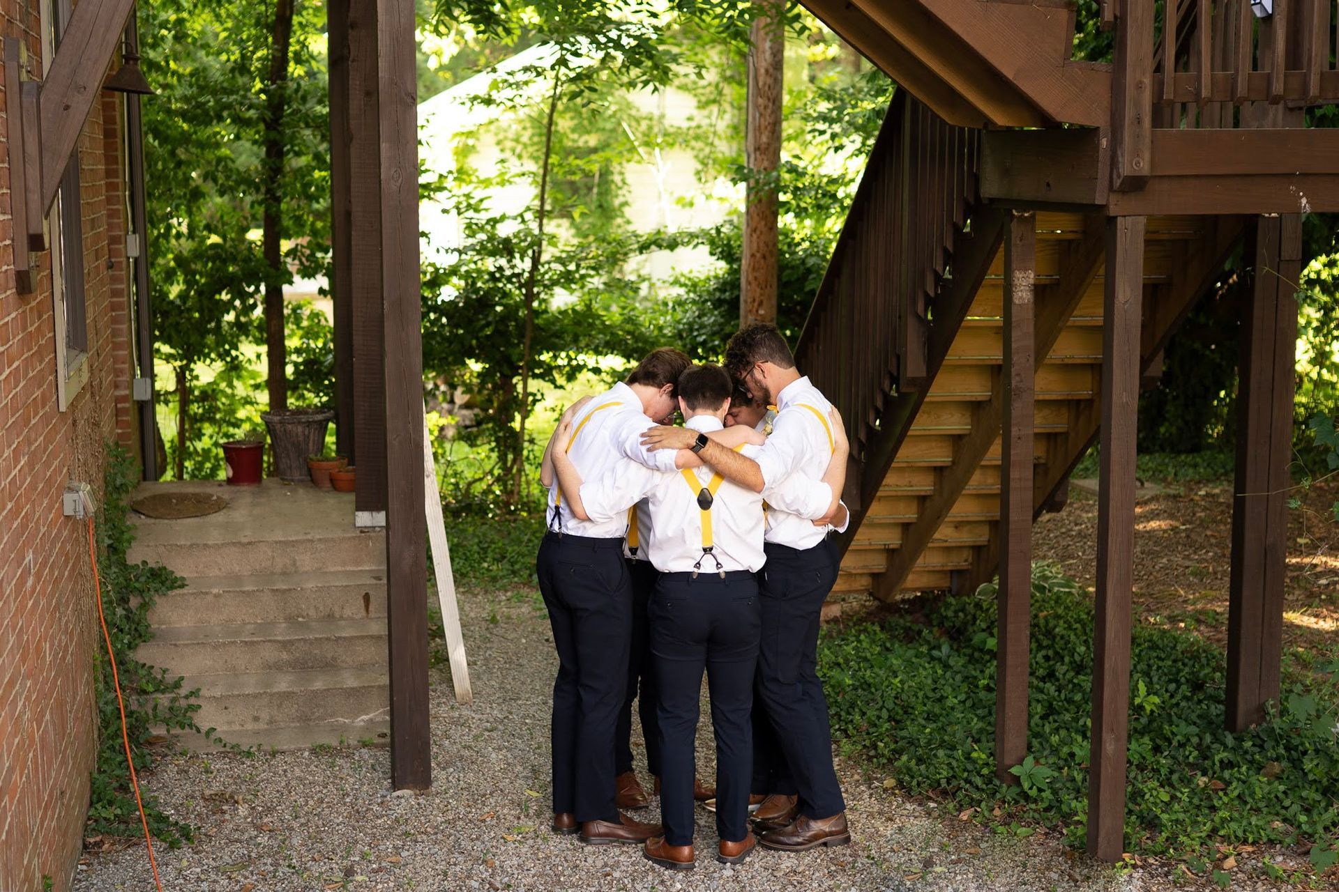 Four people in white shirts stand together outdoors under a wooden structure, heads bowed in a huddle.
