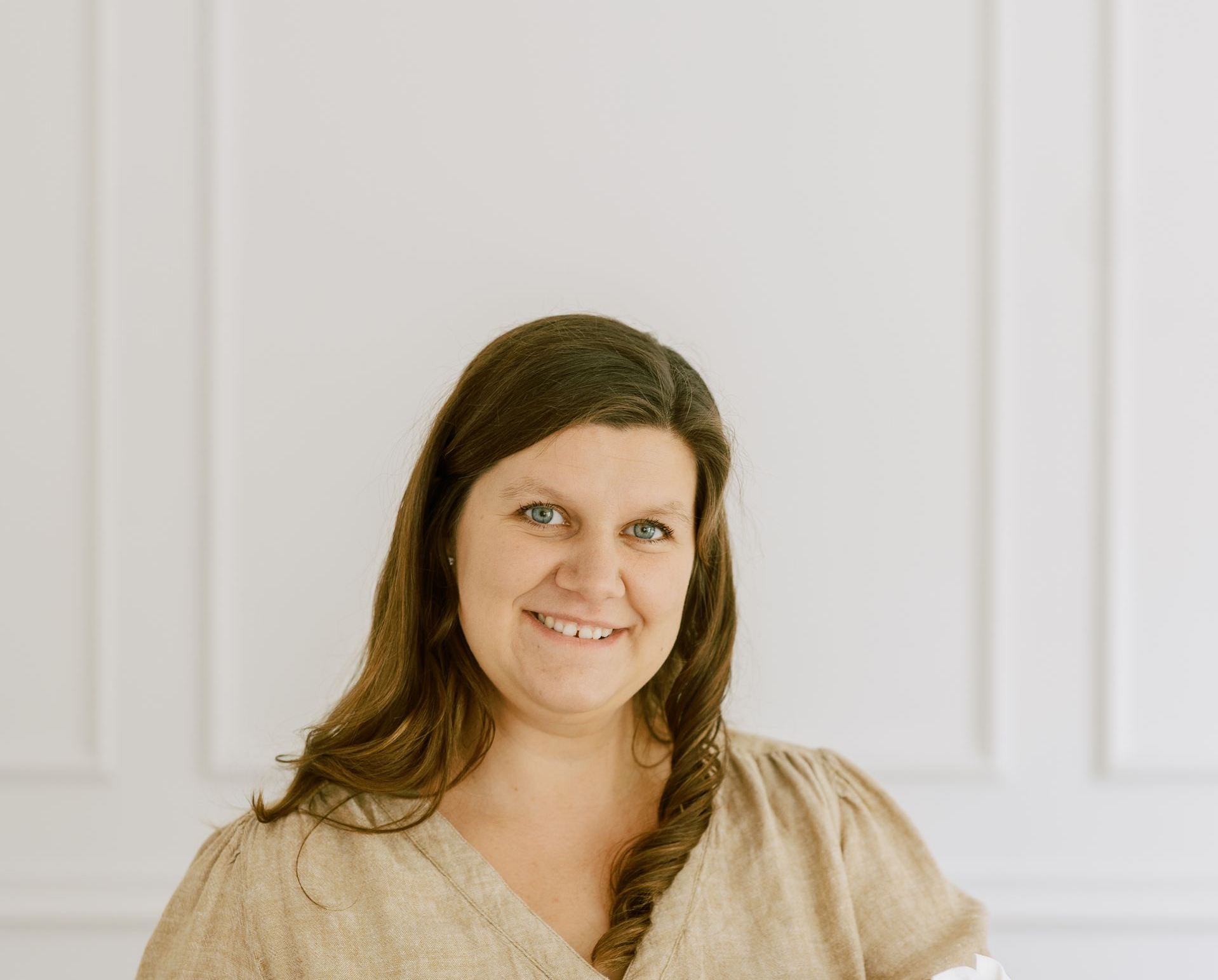Smiling woman with dark hair in a beige blouse against a light paneled wall
