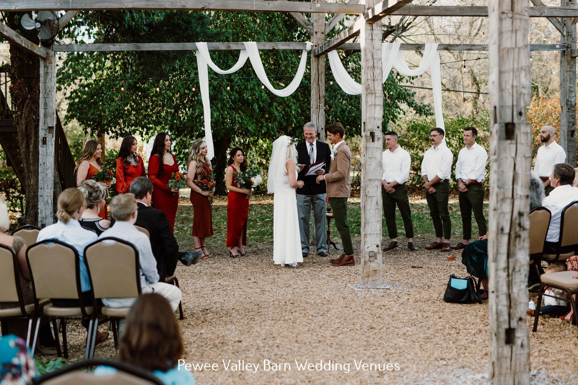 Outdoor wedding ceremony under a wooden arch, bride and groom facing an officiant with guests seated nearby