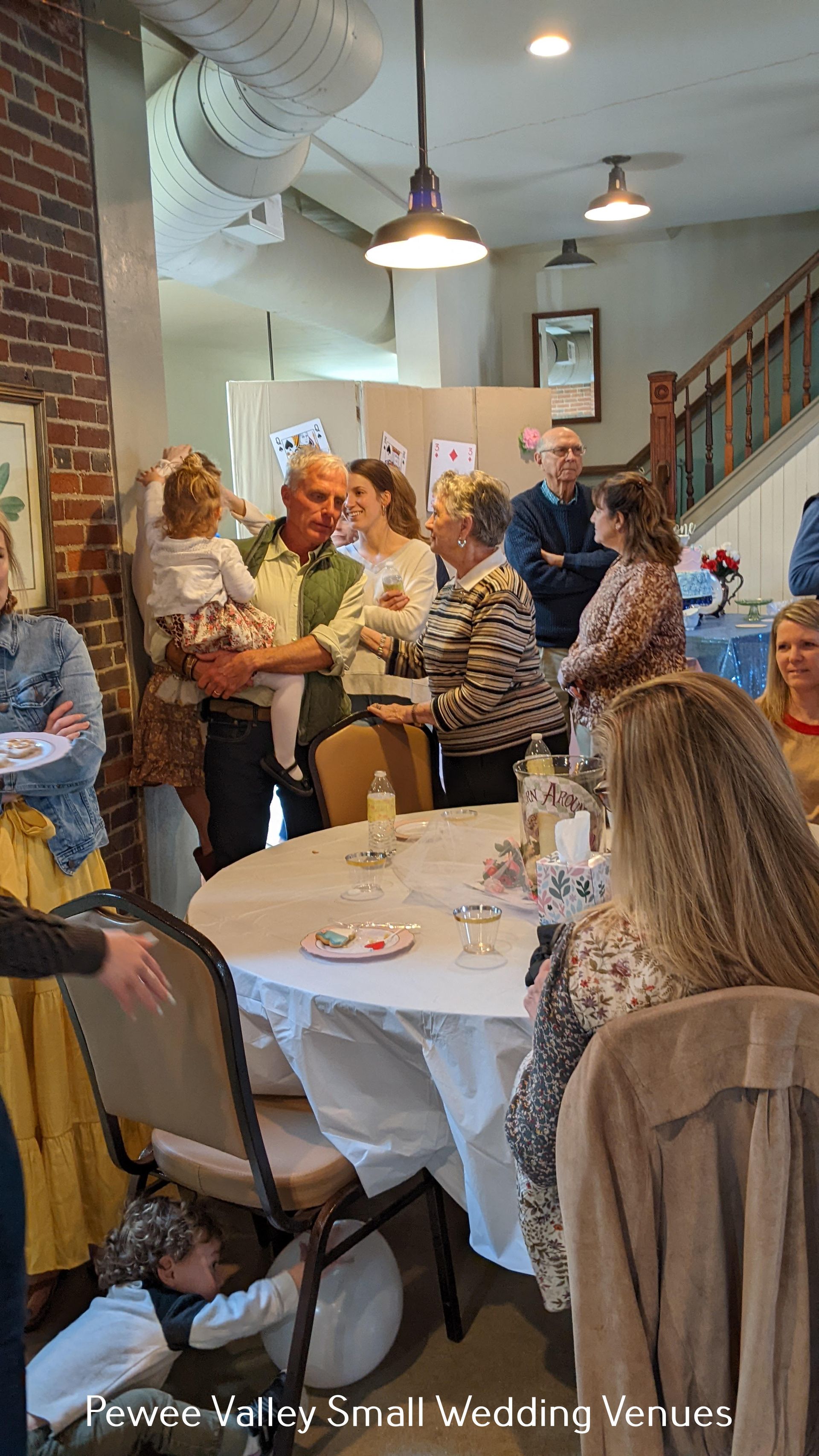 Guests mingle at a small indoor wedding reception with round tables and hanging lights.