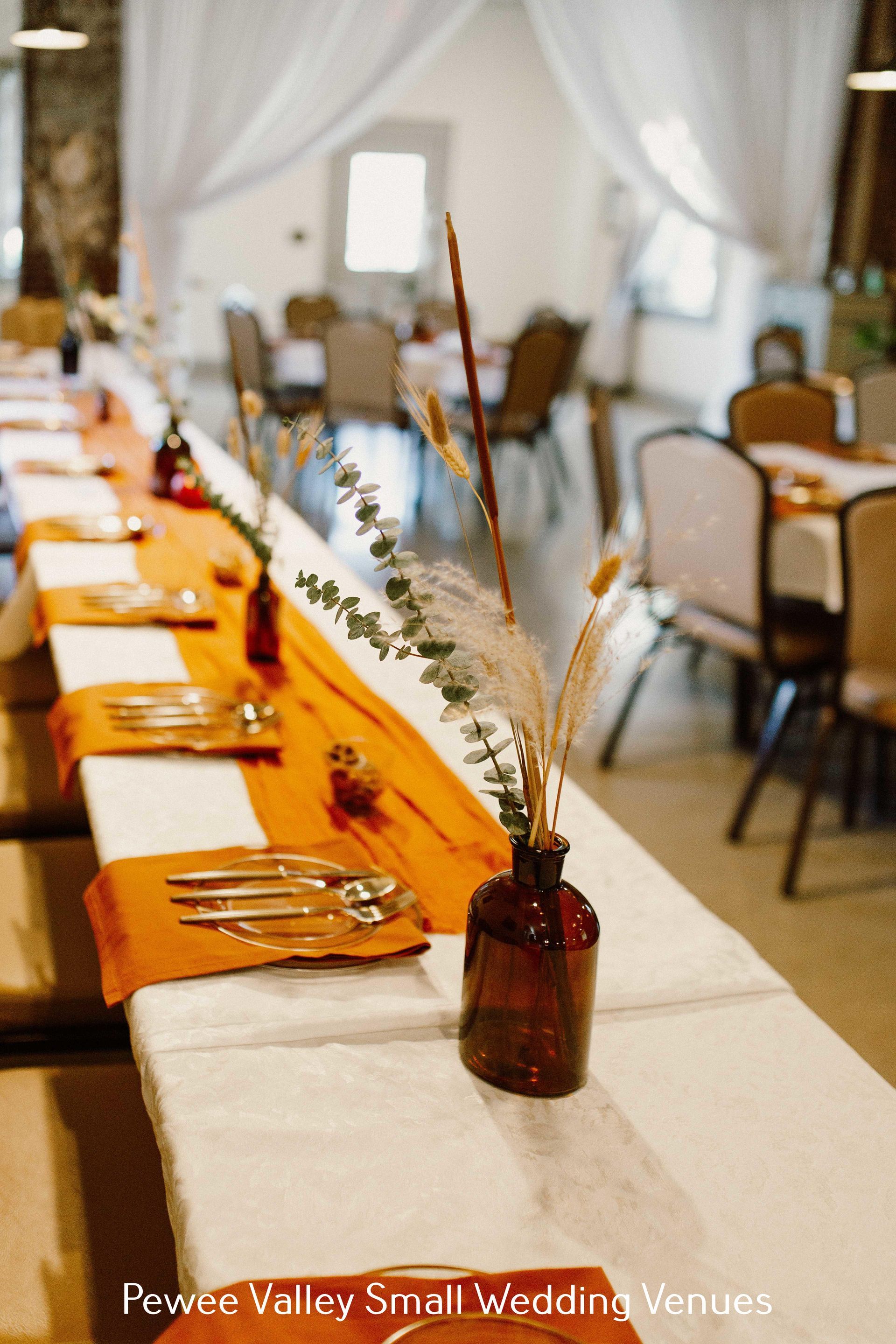 Elegant wedding reception table with orange runner, candles, and flower-filled bottles in a bright tented room