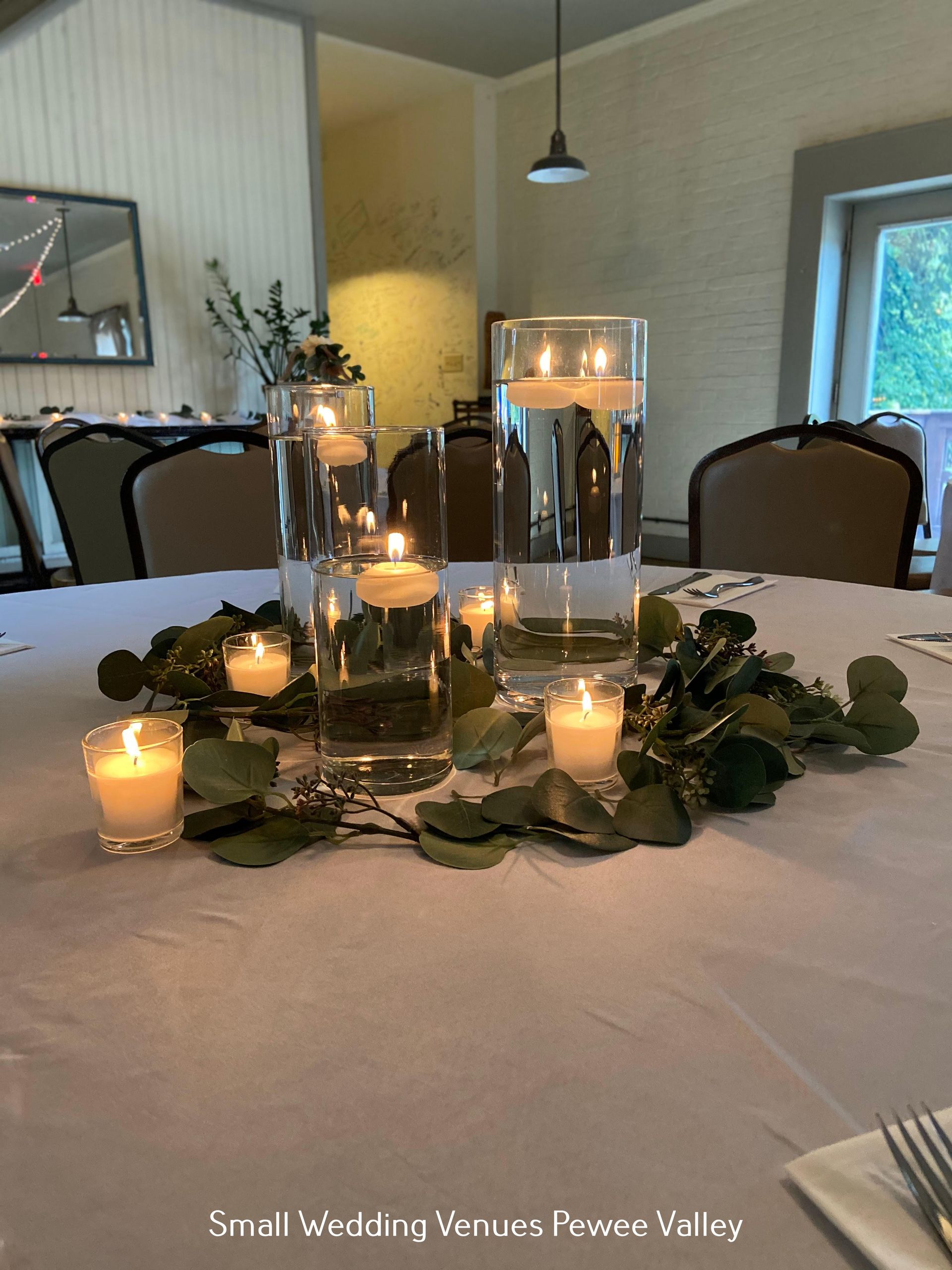 Candlelit wedding table centerpiece with greenery and glass vases in a bright venue room