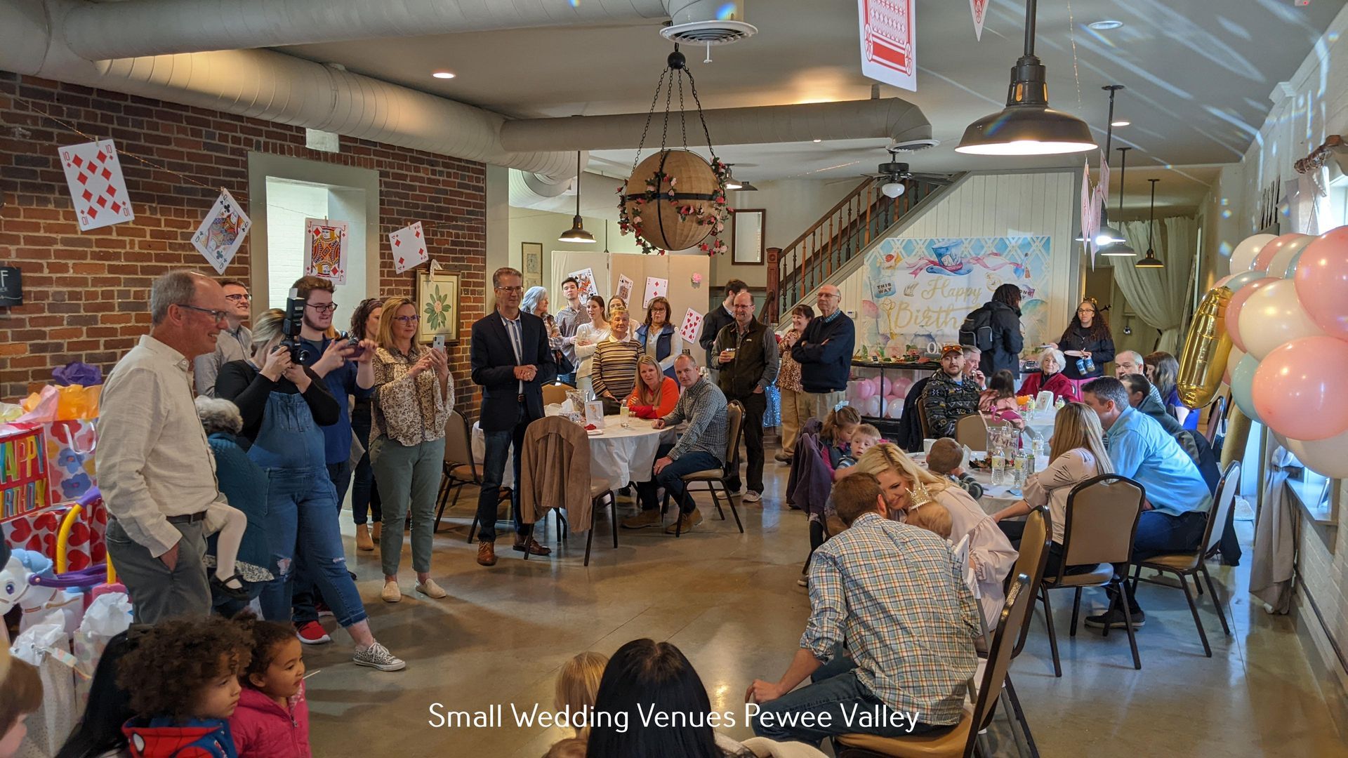 People gathered in a rustic dining hall for a social event, with balloons, tables, and hanging lights.