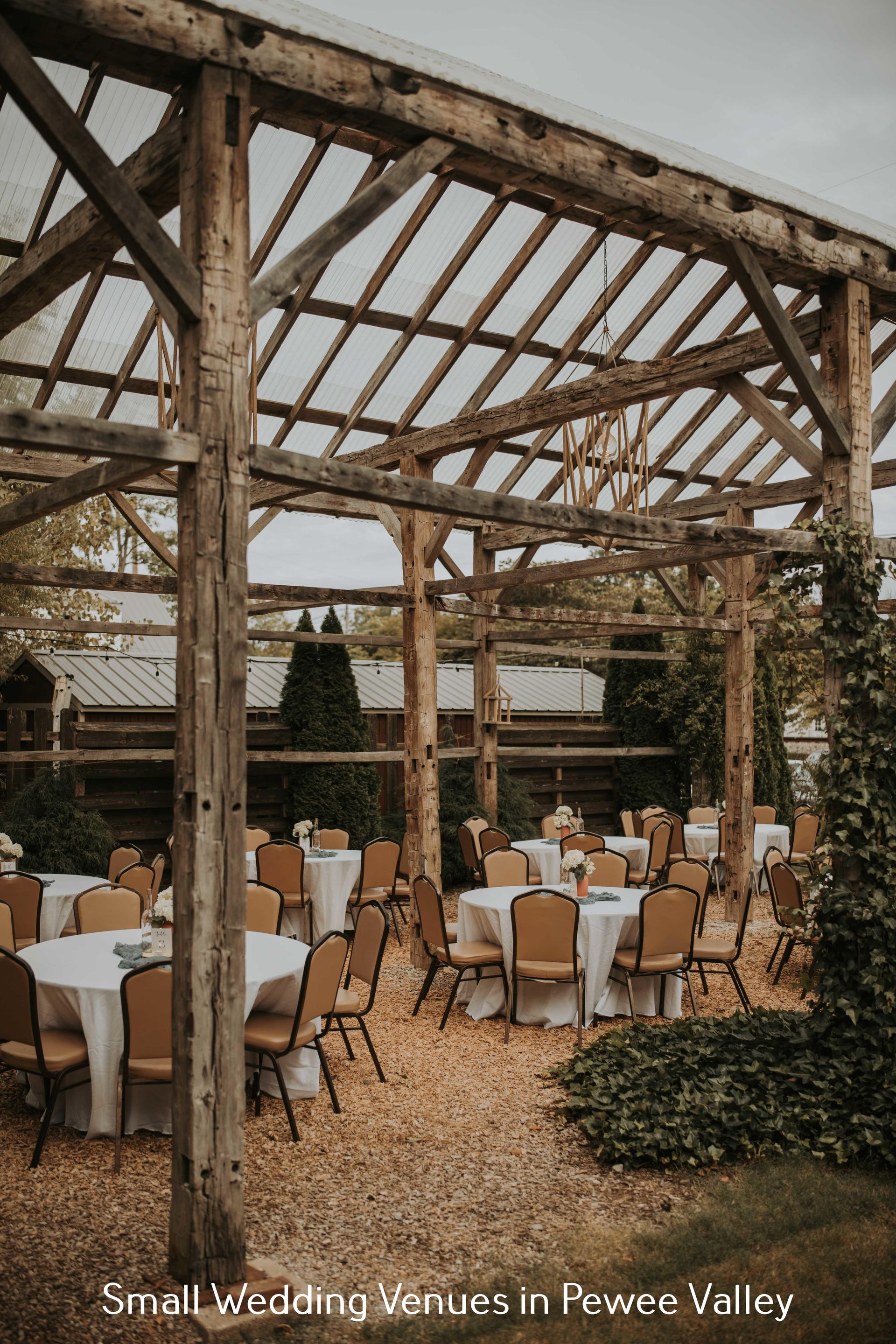 Rustic outdoor wedding reception under a wooden pergola with round tables and beige chairs in a garden courtyard