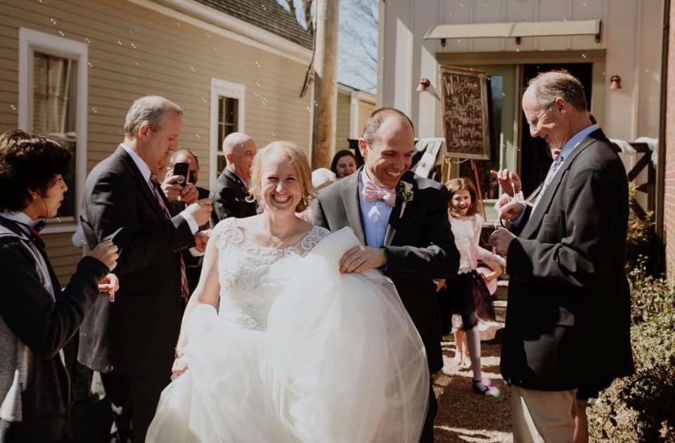 Wedding couple exiting venue with guests applauding, the bride in a white gown and groom in a dark suit.