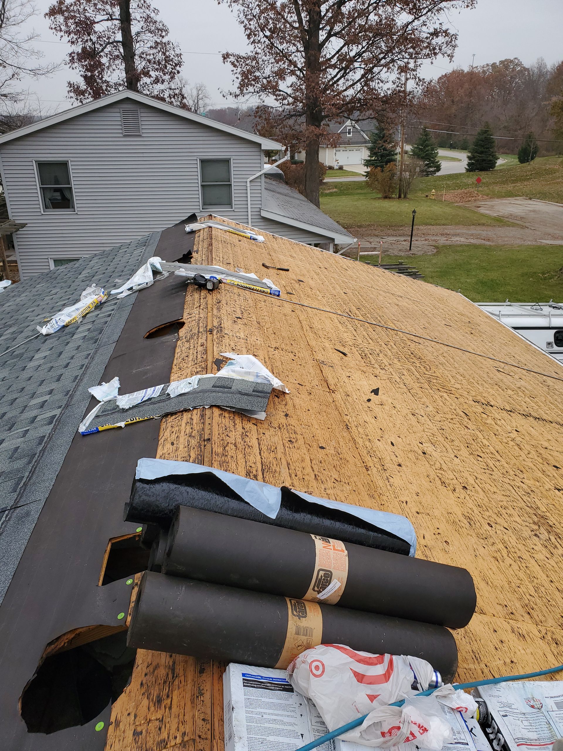Roof in progress: partially covered with shingles, exposed wooden boards, and roofing materials. Overcast day.