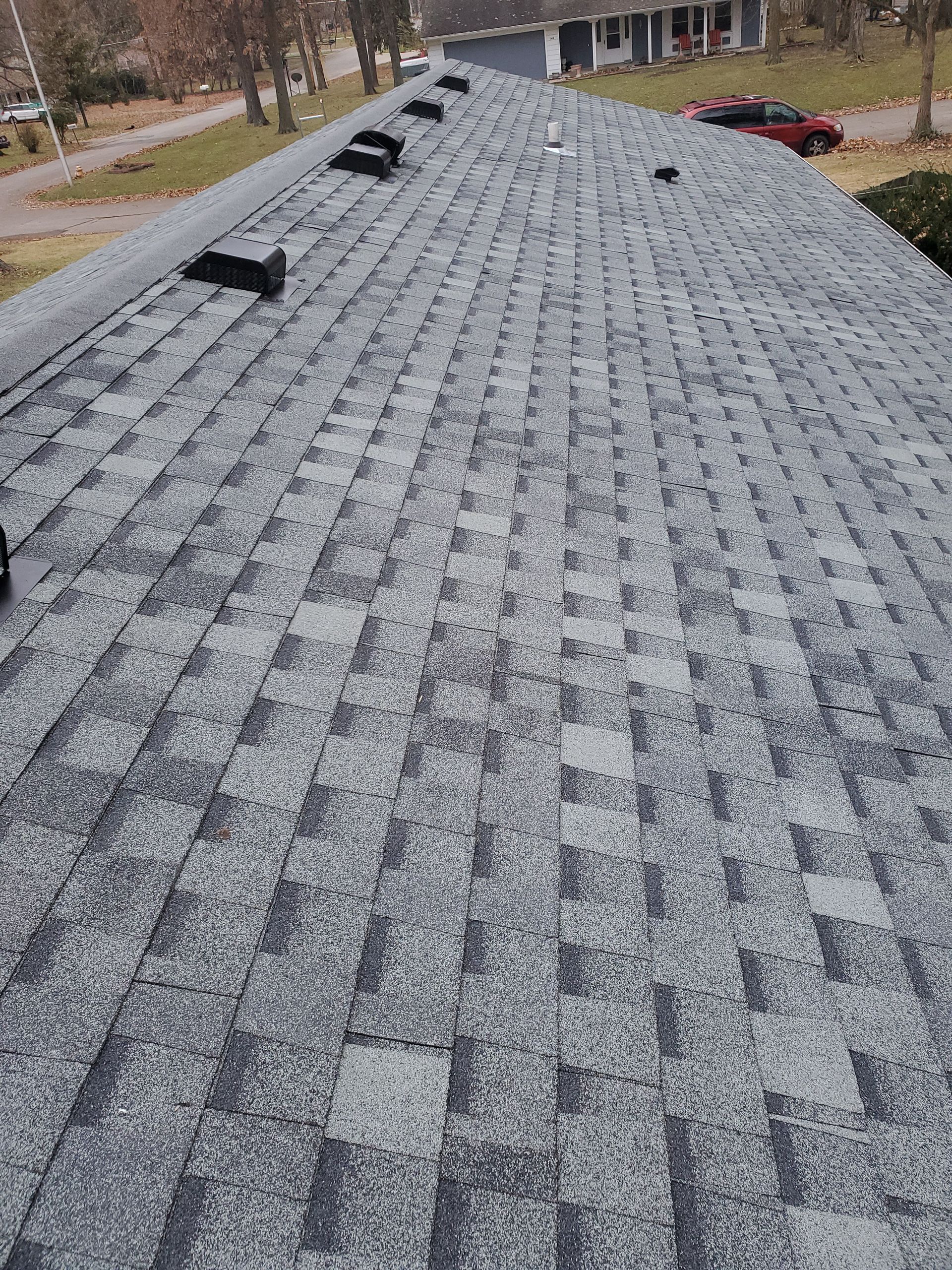 Gray asphalt shingle roof on a house, with black vents along the ridge and a glimpse of neighborhood in the background.