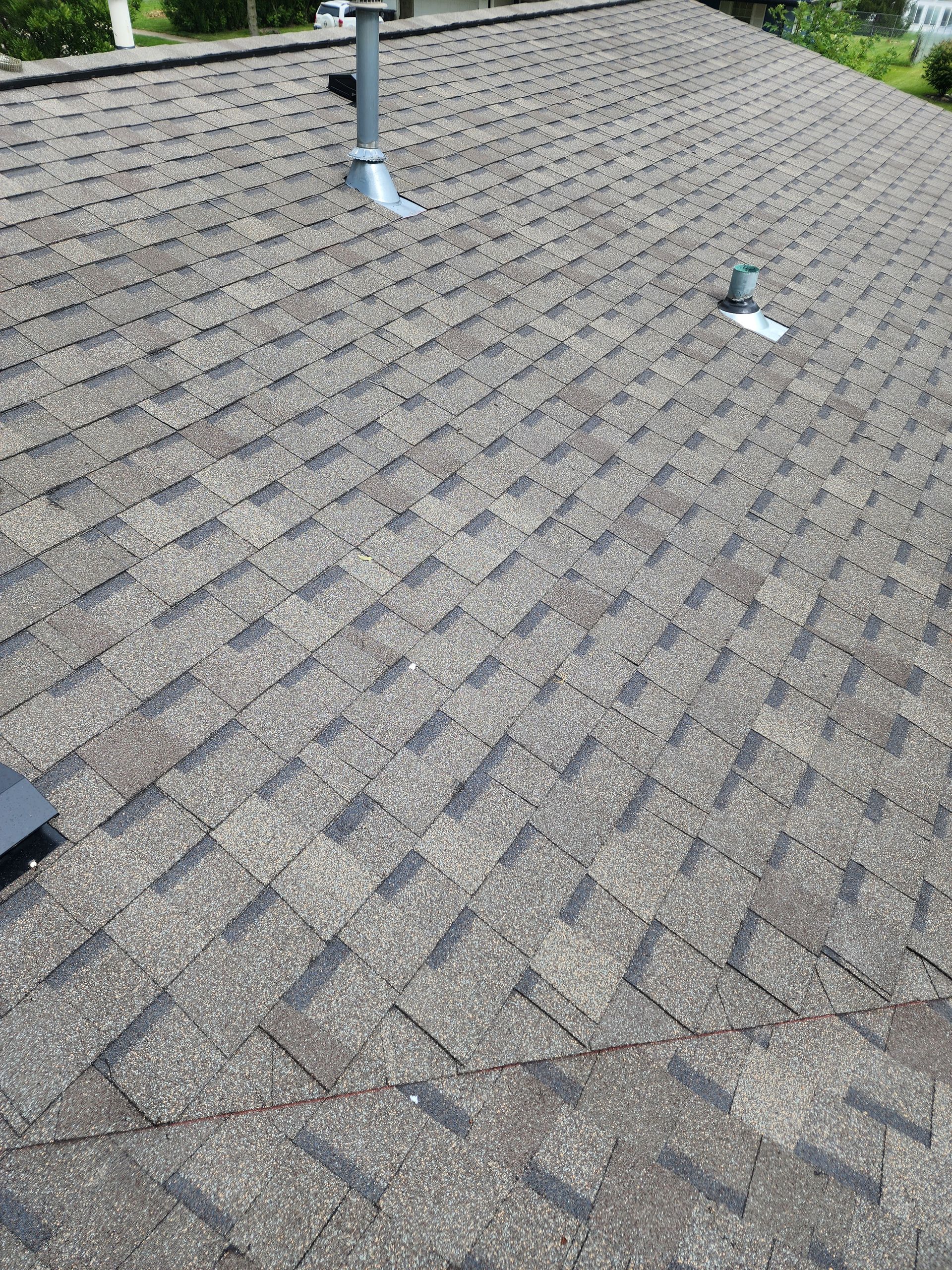Gray asphalt shingle roof with two vents and a chimney against a background of foliage.