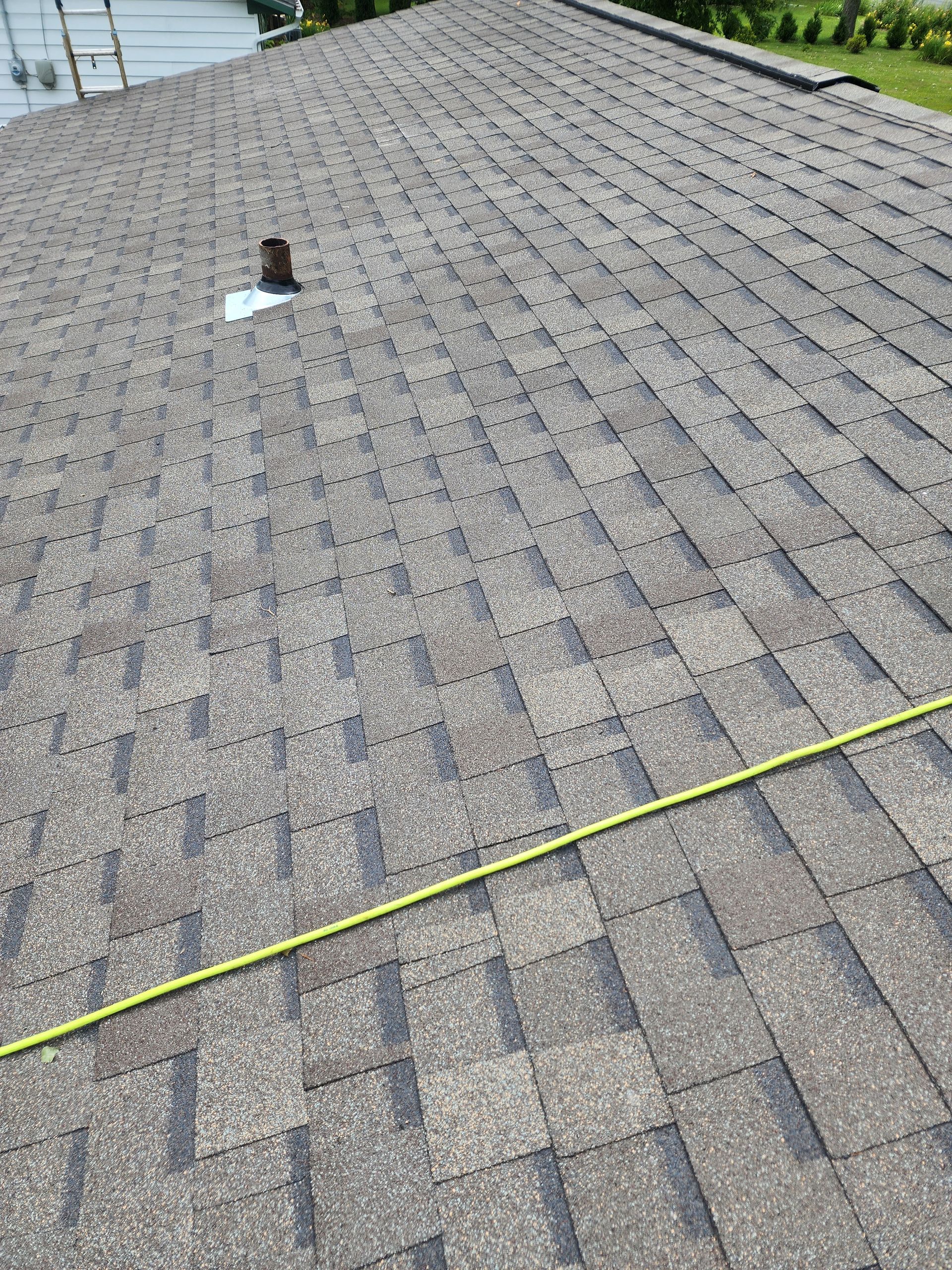 Close-up view of a brown asphalt shingle roof with a vent pipe. A yellow measuring tape is on the roof.